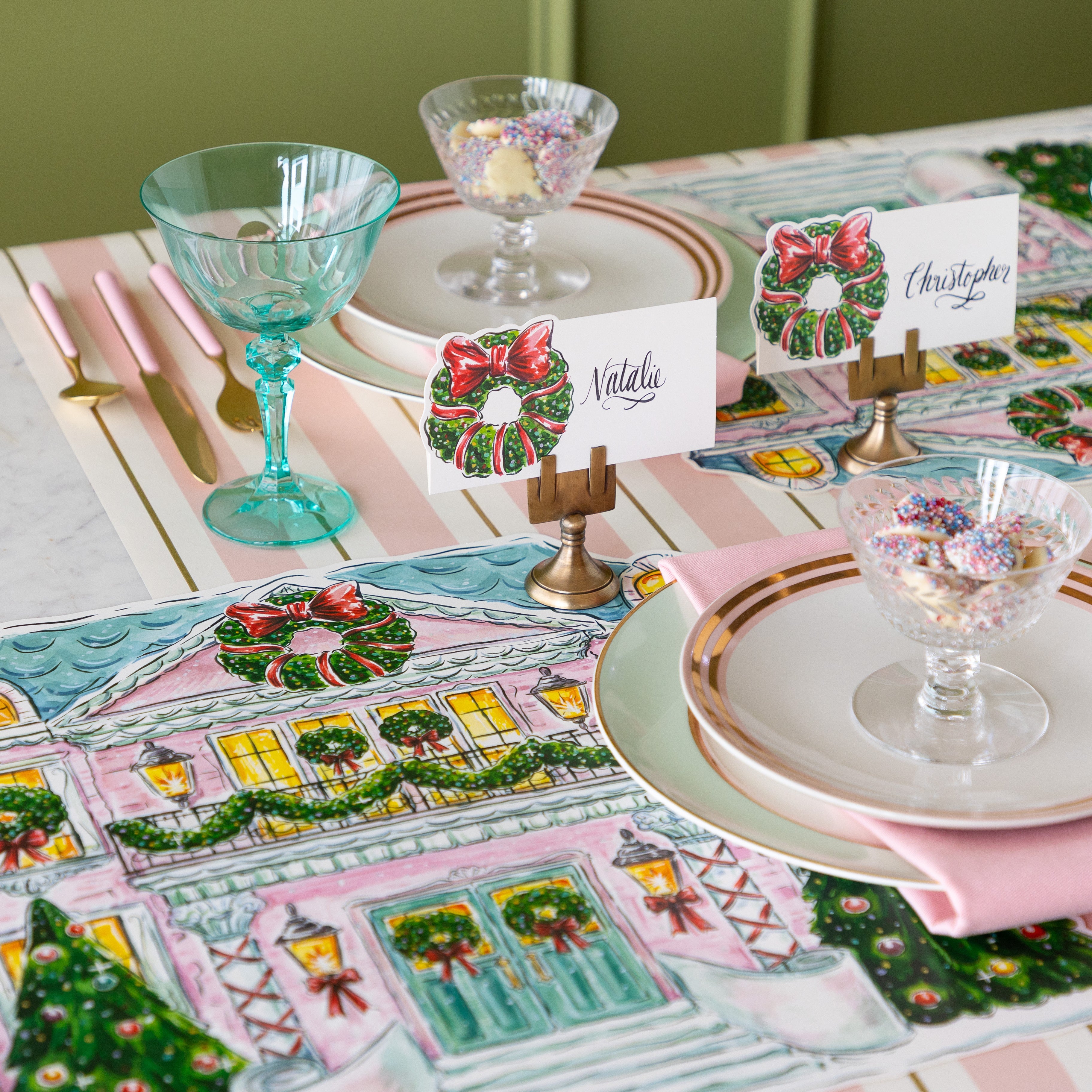 A place setting featuring the Holiday Wreath Place Cards held in the Brass Place Card Holders, plates with a pink napkin in between and bowl of white chocolate buttons atop, on the Pink & Gold Awning Stripe Runner.