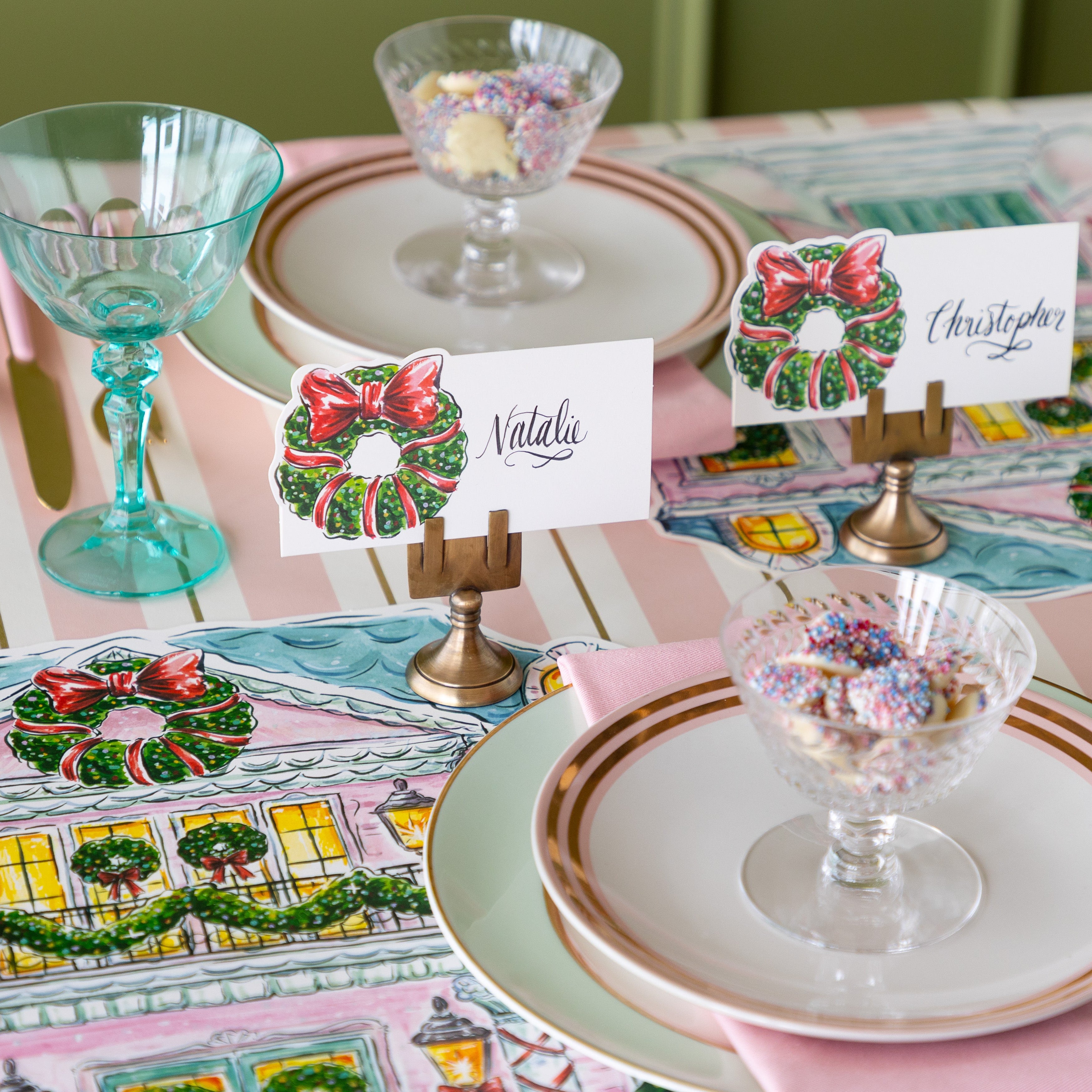 A table setting featuring the Holiday Wreath Place Cards held in the Brass Place Card Holders, Die-cut Home for the Holidays Placemats, seafoam and gold rimmed plates, pink napkins, and bowls of white chocolate buttons on the Pink & Gold Awning Stripe Runner.