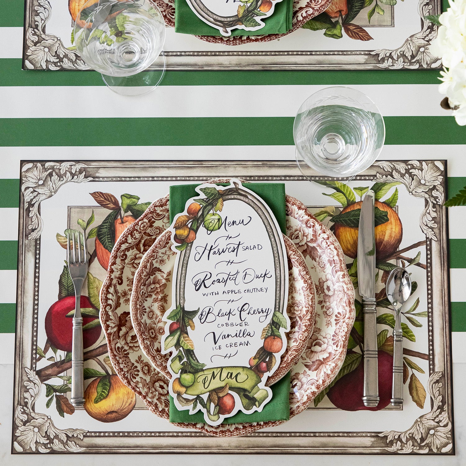 A Heirloom Apples Table Card with a menu written inside the frame and &quot;Mac&quot; written on the ribbon, resting on the plate of an elegant place setting, from above.