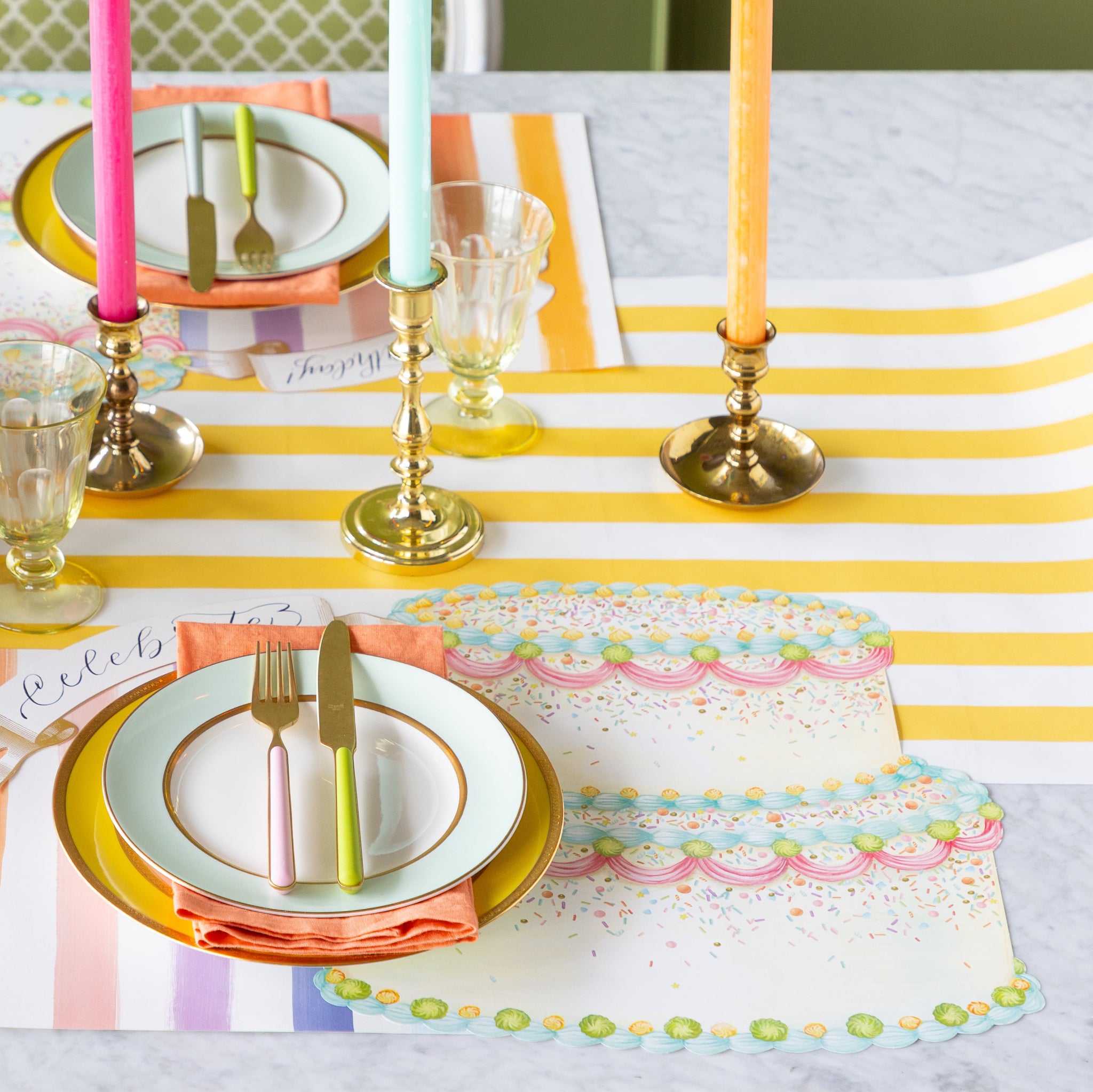 A colorful table setting featuring the Die-cut Birthday Cake Placemat overlapping the Sorbet Painted Stripe Placemat, colorful dinnerware and Classic Gold Banner Table Accent on the Marigold Classic Stripe Runner.