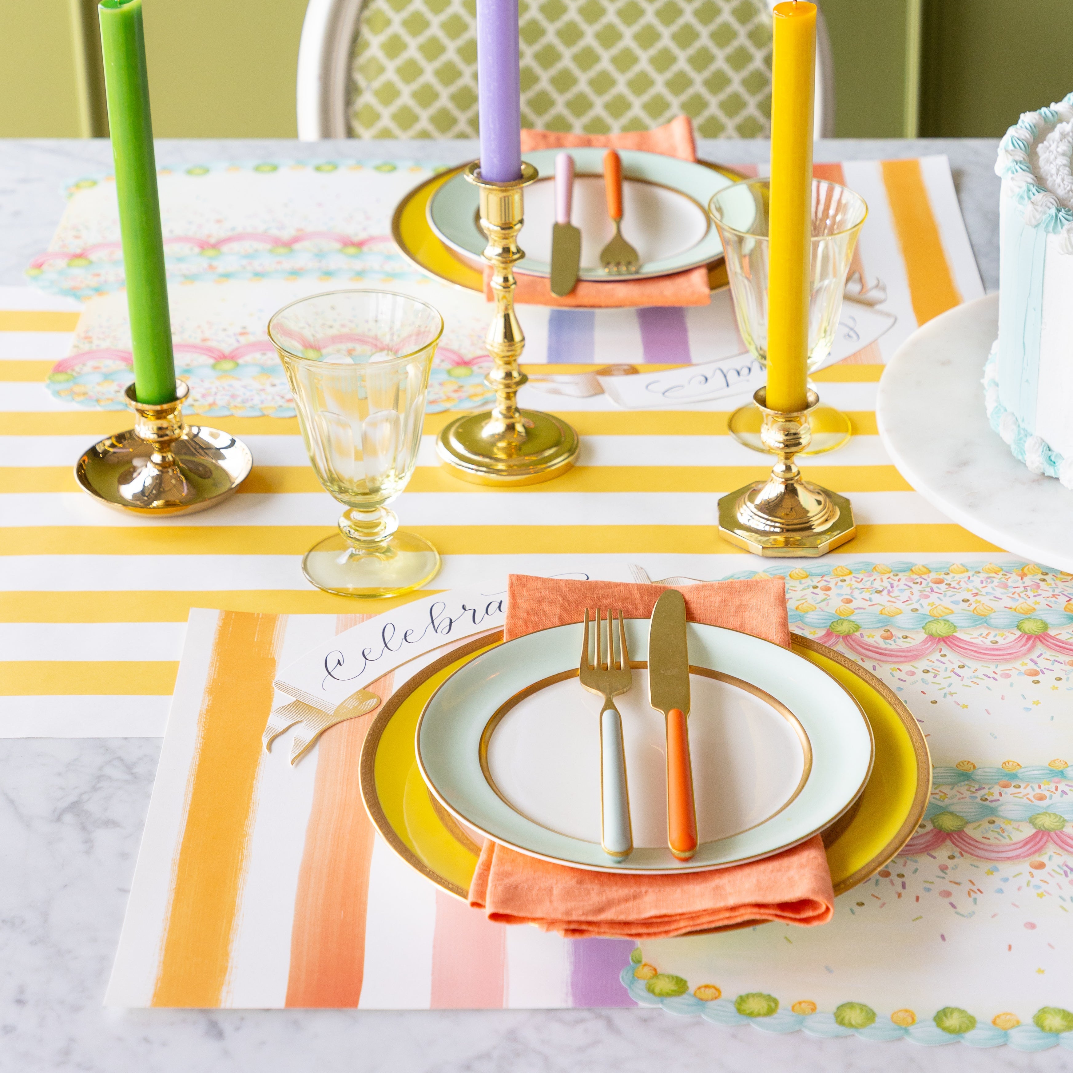 A colorful table setting featuring the Die-cut Birthday Cake Placemat overlapping the Sorbet Painted Stripe Placemat, colorful dinnerware and Classic Gold Banner Table Accent on the Marigold Classic Stripe Runner.