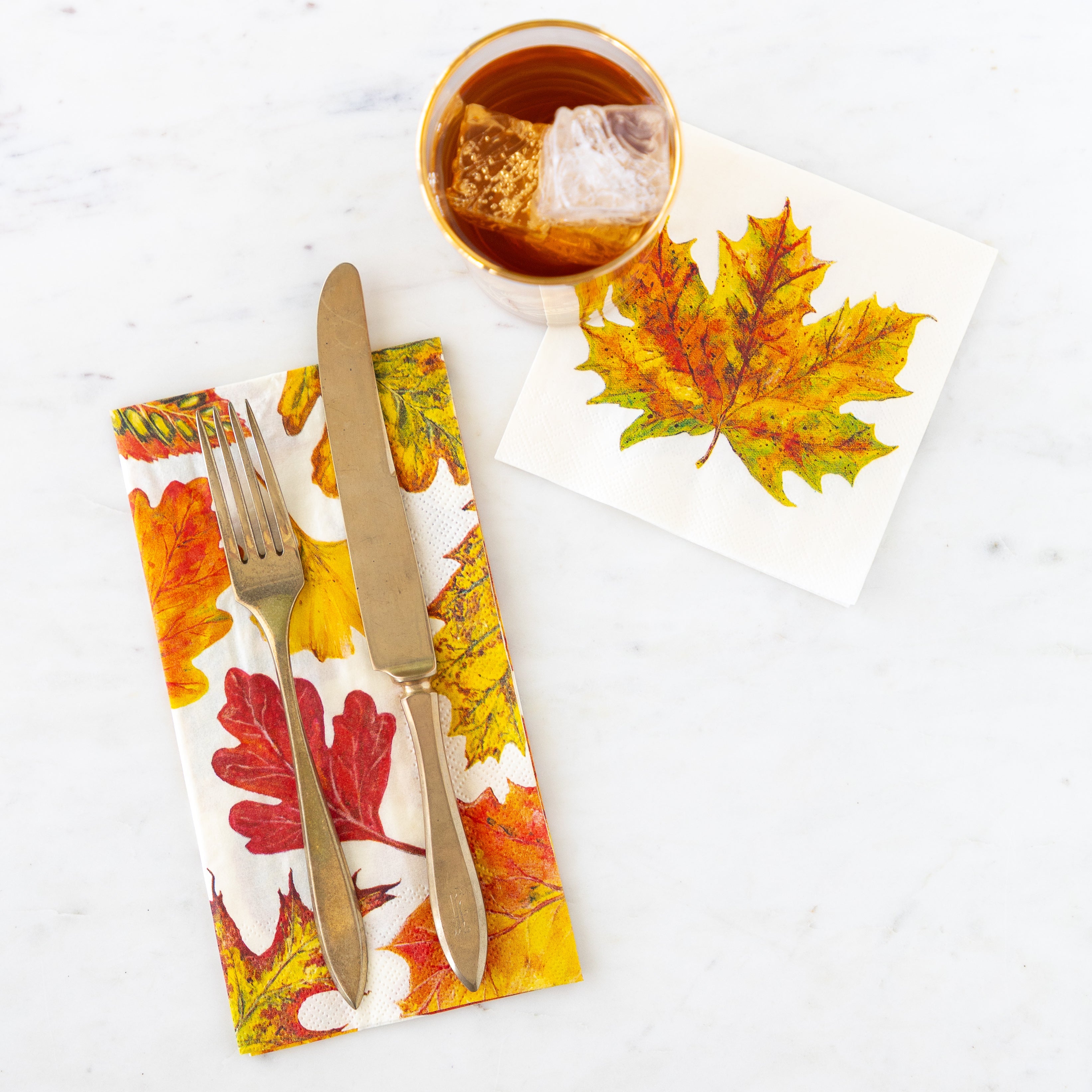 Fall Foliage Guest Napkin with a fork and knife on top, next to the Fall Foliage Cocktail Napkin with a glass of tea slightly covering the napkin.