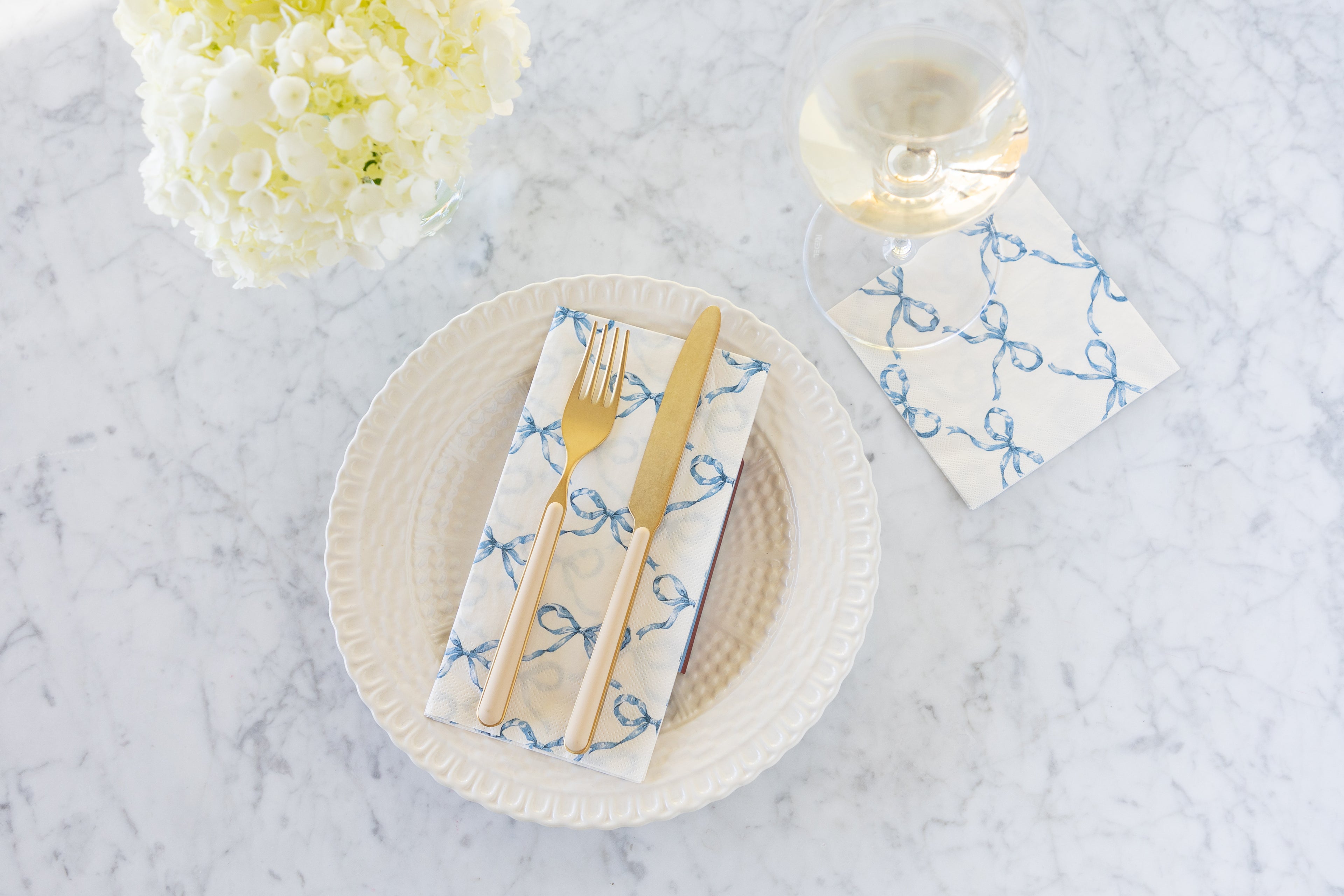 A plate with the Heirloom Blue Bow Lattice Guest Napkin and gold/peach fork and knife atop, flowers, a glass of wine and Heirloom Blue Bow Cocktail Napkin underneath.
