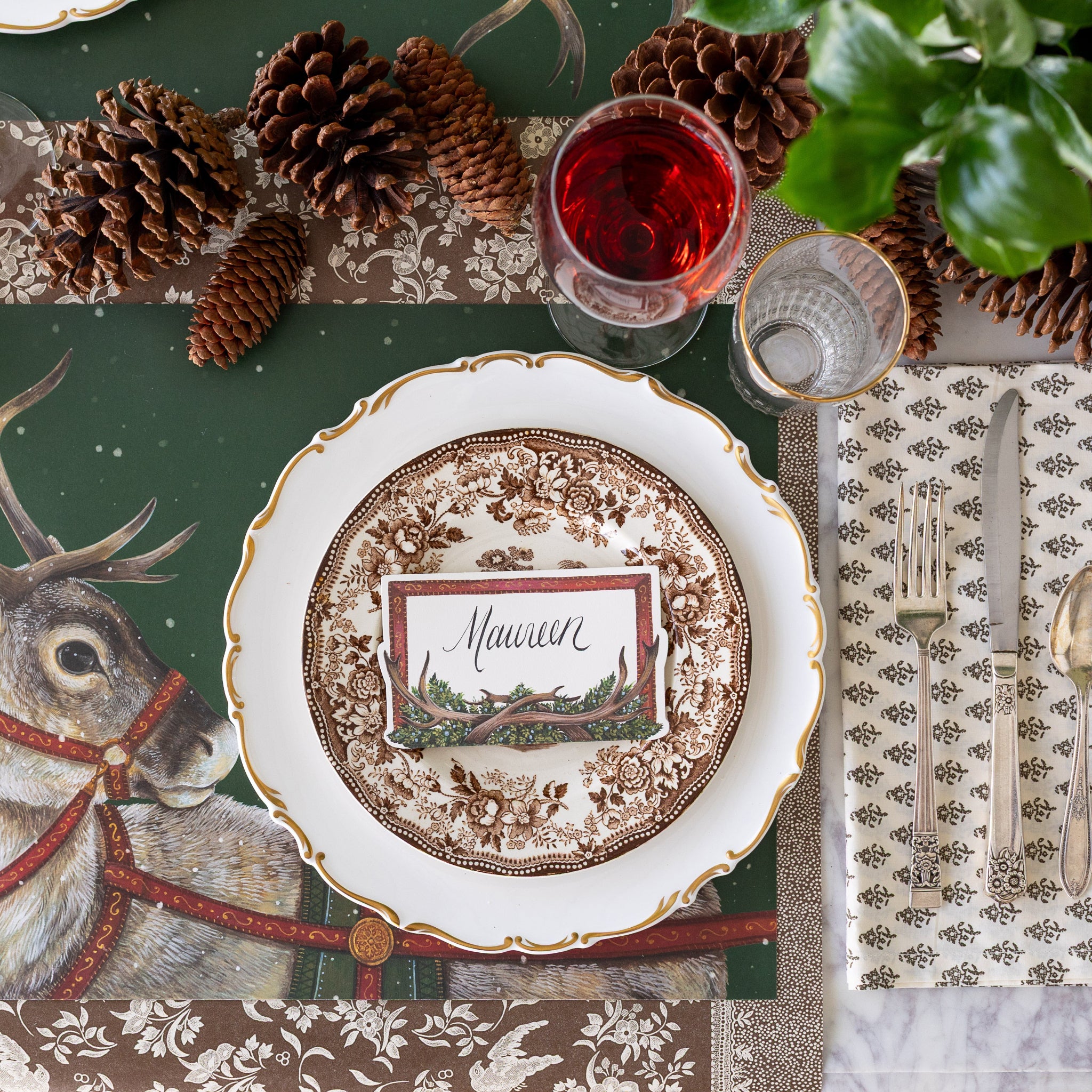 Christmas themed table setting featuring the Antler Frame Place Card atop dinner plates, Holiday Reindeer Placemat underneath, pinecones and floral napkin on the Brown Regal Peacock Runner.