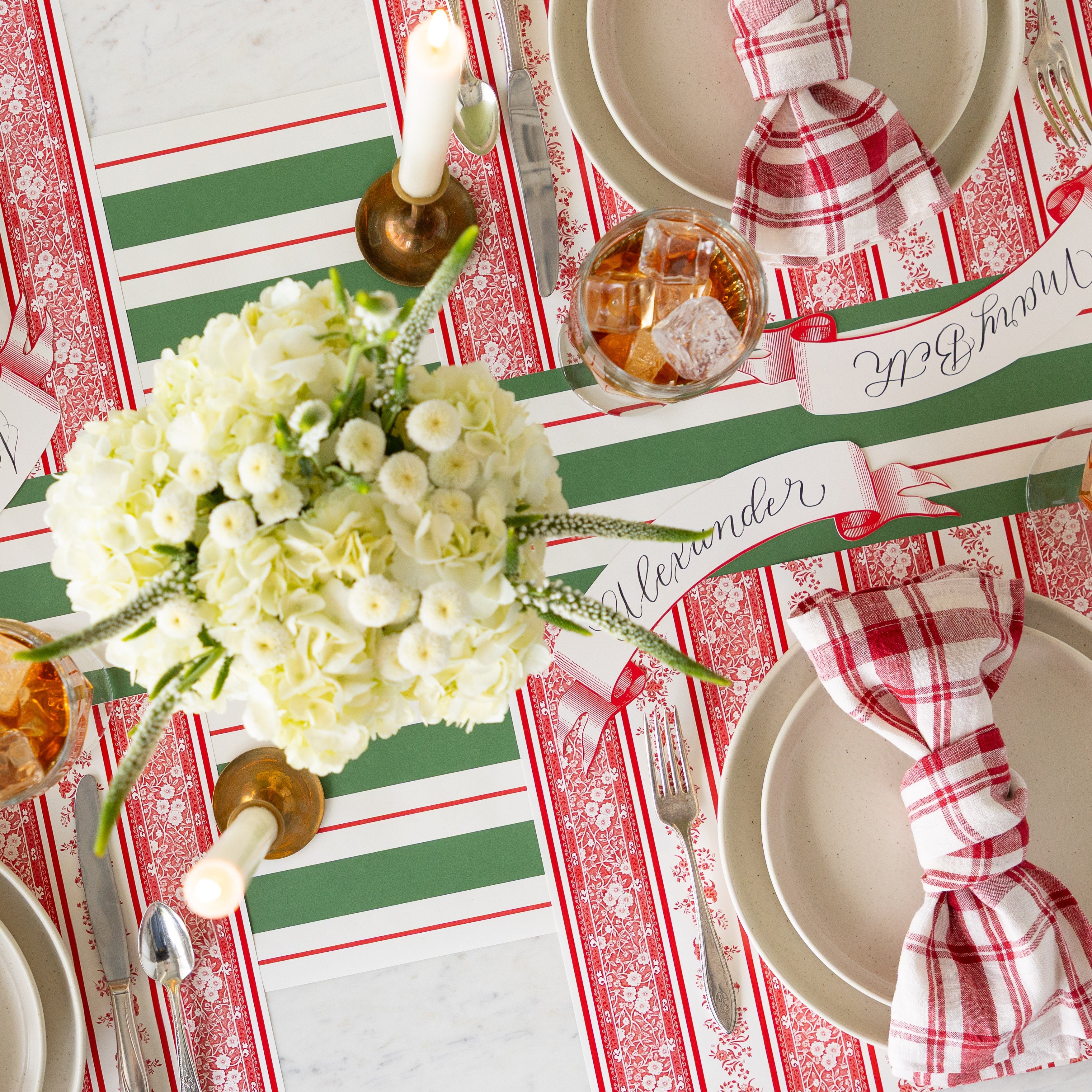 An overhead shot of a table setting with a floral centerpiece and candles, featuring the Red Burleigh Stripe Placemats, dinner plates with red and white checkered napkins atop, vintage flatware, Red Classic Banner Table Accents and Green & Red Awning Stripe Runner underneath.
