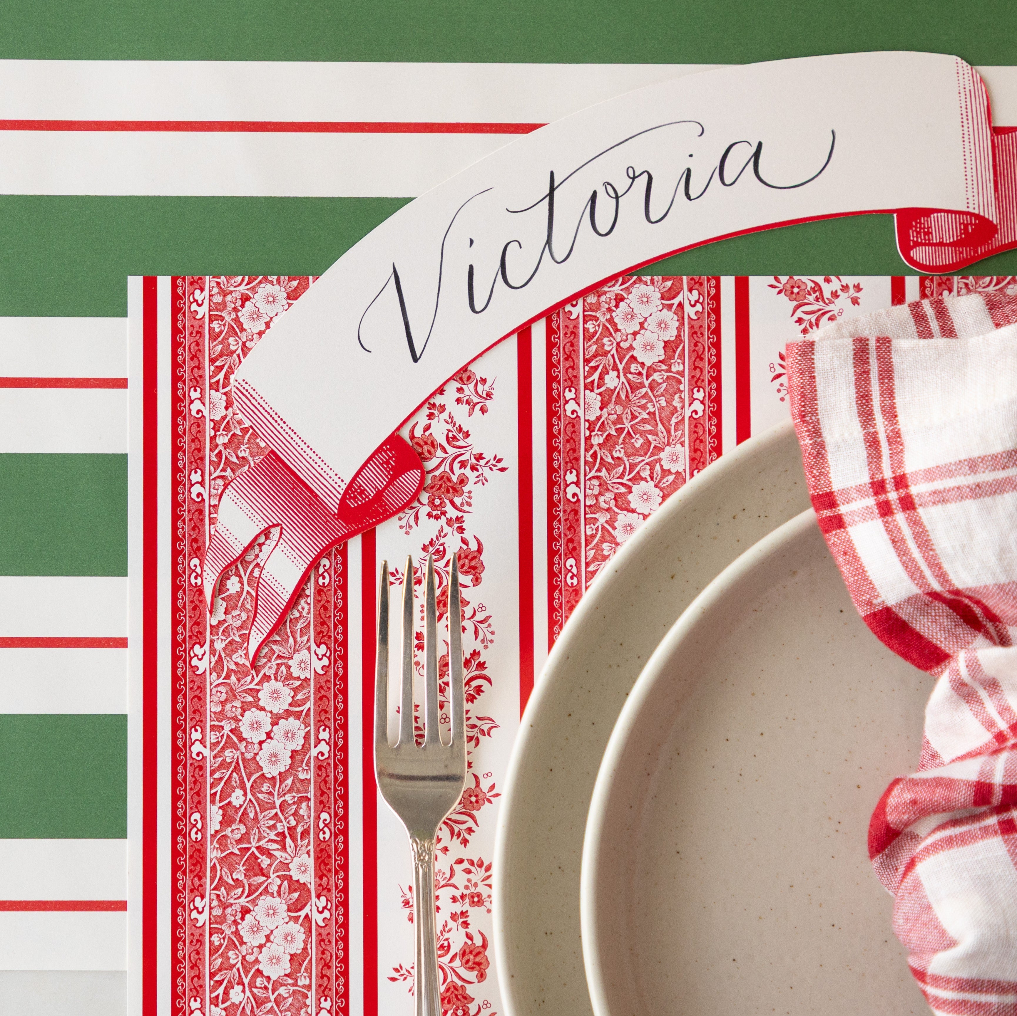 The Red Burleigh Stripe Placemat under a place setting with white dinnerware, a red and white striped napkin, a vintage fork, Classic Red Banner Table Accent reading "Victoria" and Green & Red Awning Stripe Runner underneath.