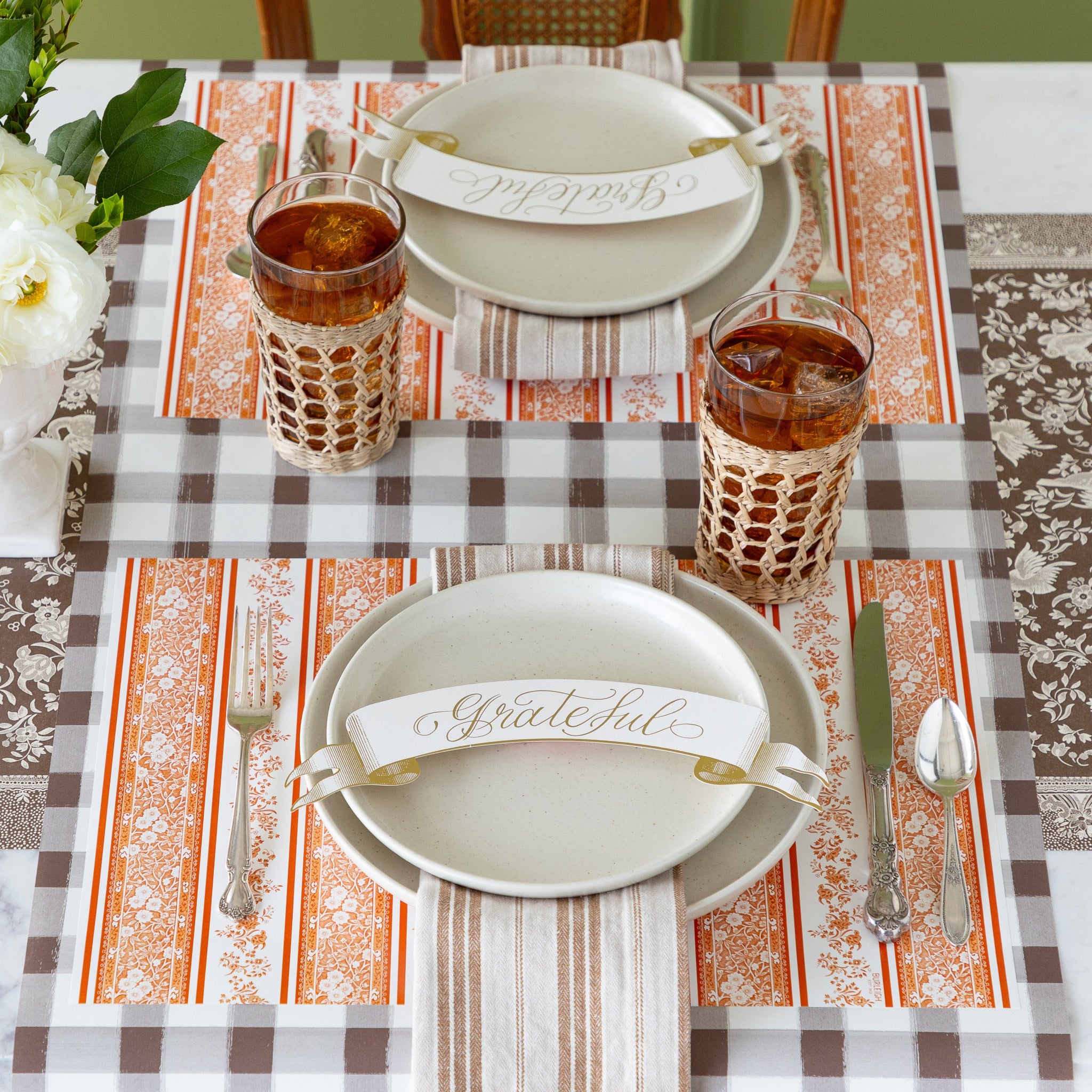 A table setting featuring the Pumpkin Burleigh Stripe Placemats with vintage silverware, vanilla dinnerware, tan striped napkins, Grateful Banner Table Accents and a SeagrassCaged Tumbler with iced tea atop, on the Brown Painted Check Runner.