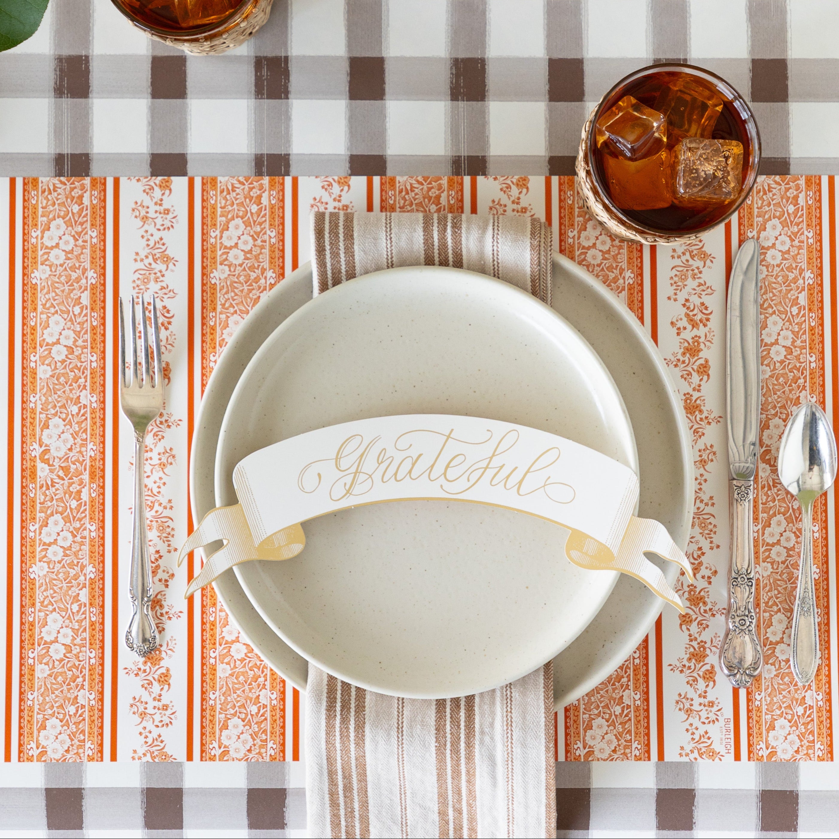 A place setting featuring the Pumpkin Burleigh Stripe Placemat with vintage silverware, vanilla dinnerware, a tan striped napkin and Grateful Banner Table Accent atop, on the Brown Painted Check Runner.