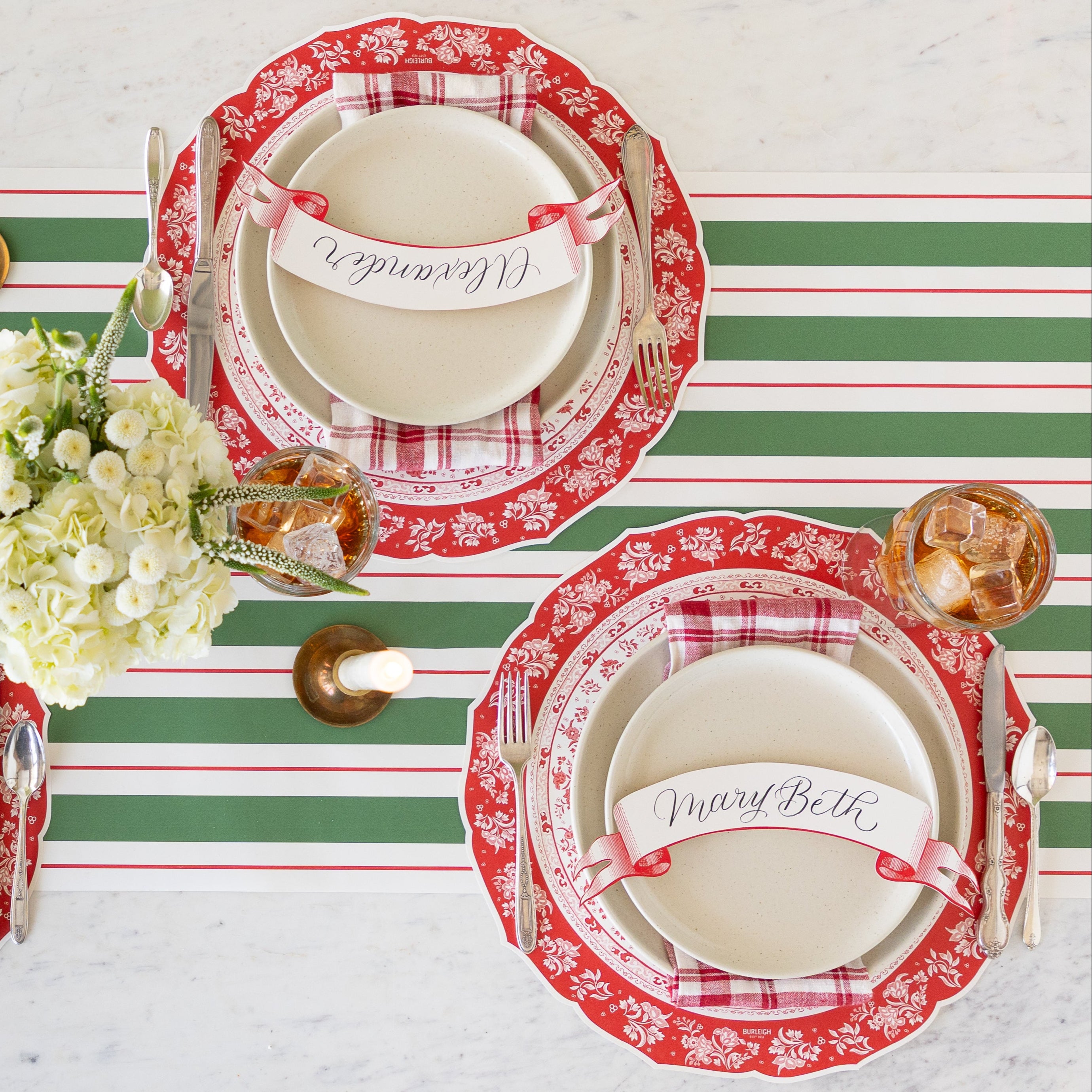 A table setting with a floral centerpiece and a lit candle, featuring the Die-cut Red Regal Peacock Placemat, off-white dinner plates with a red-and-white checkered napkin in between and Classic Red Banner Table Accent atop, vintage flatware, glasses of tea, and Green & Red Awning Stripe Runner underneath.