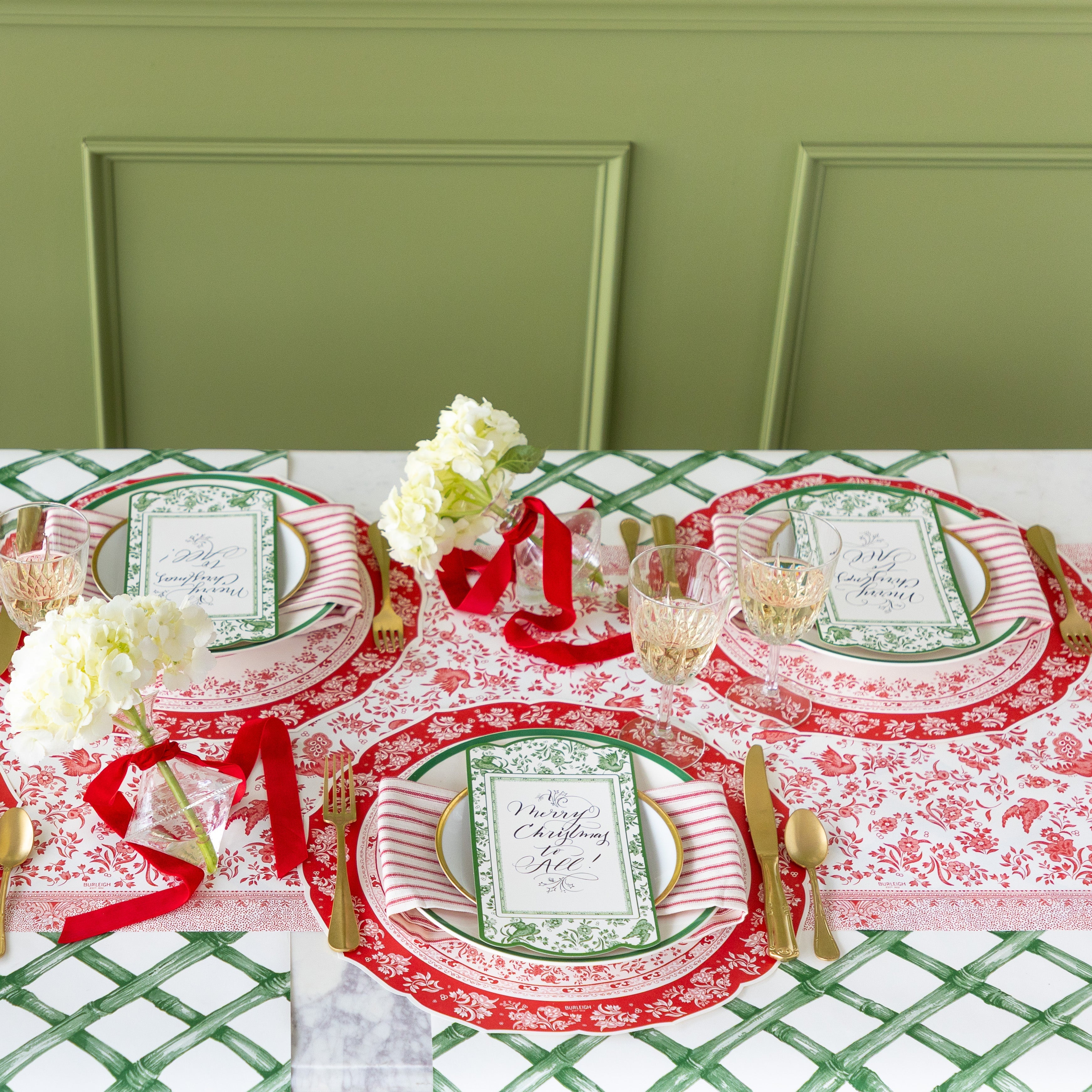 A table for three is set featuring the Die-cut Red Regal Placemat, dinner plates with a red-and-white striped napkin in between, Green Regal Peacock Table Accent reading "Merry Christmas to All!" atop, gold flatware, Green Lattice Runner rolled out vertically underneath and Red Burleigh Peacock Runner rolled out horizontally underneath.