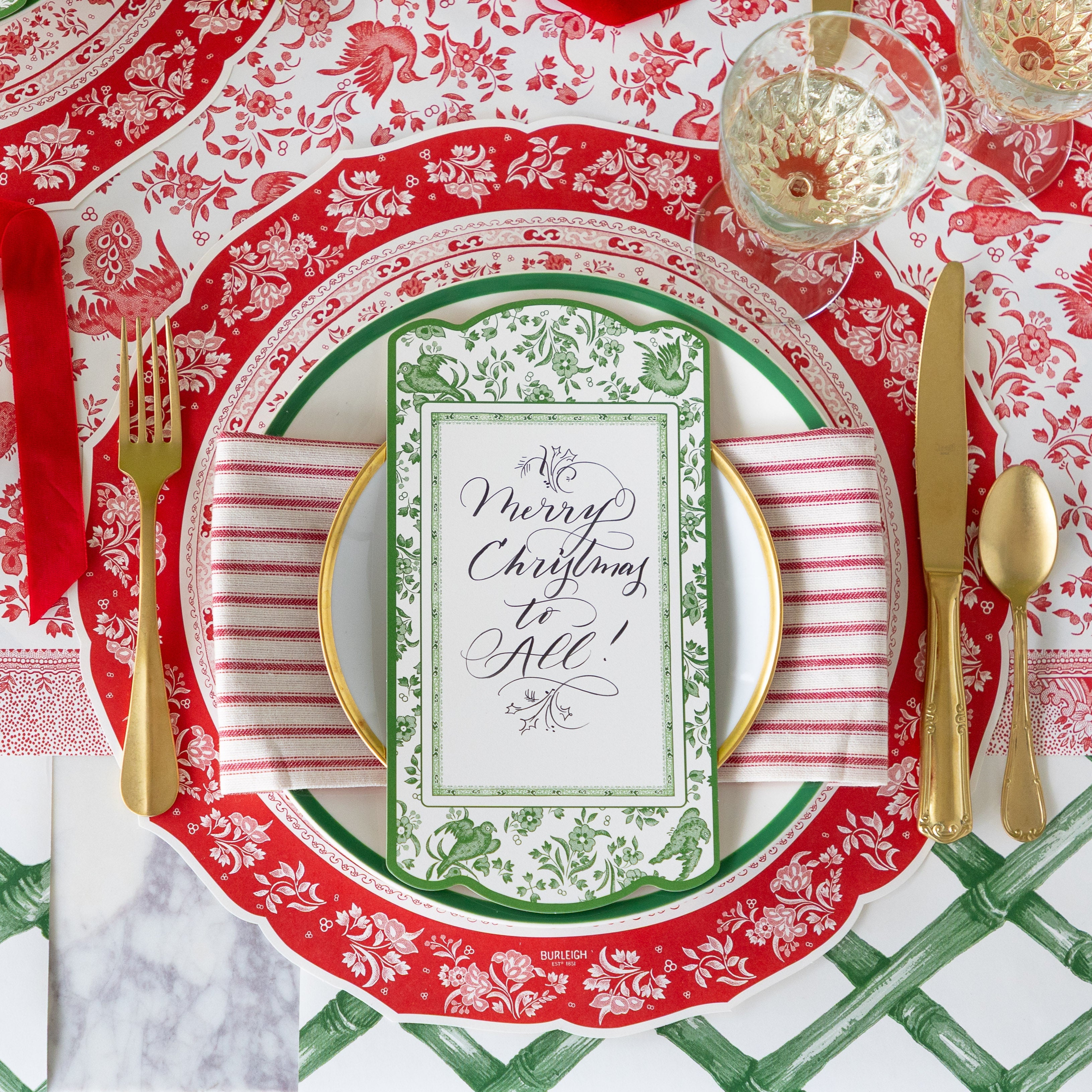An elegant place setting featuring the Die-cut Red Regal Placemat, green and gold rimmed plates with a red-and-white striped napkin in between, Green Regal Peacock Table Accent reading "Merry Christmas to All!" atop, gold flatware, and Green Lattice Runner and Red Burleigh Peacock Runner underneath.