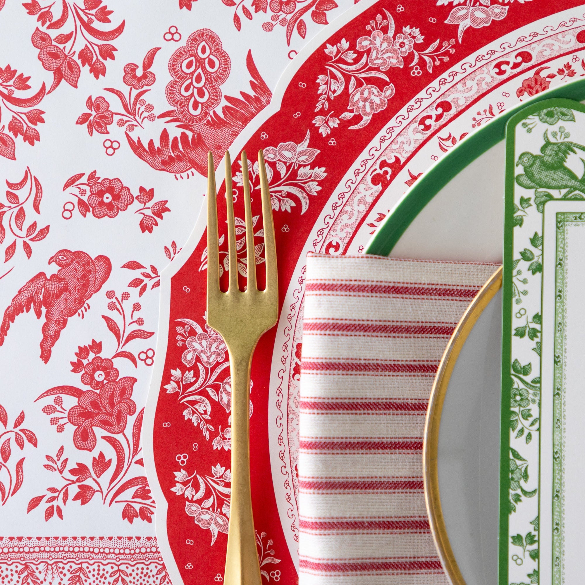 An elegant place setting featuring the Die-cut Red Burleigh Placemat, a gold fork, green-rimmed dinner and gold-rimmed salad plate, red-and-white striped napkin and Red Regal Peacock Runner underneath.