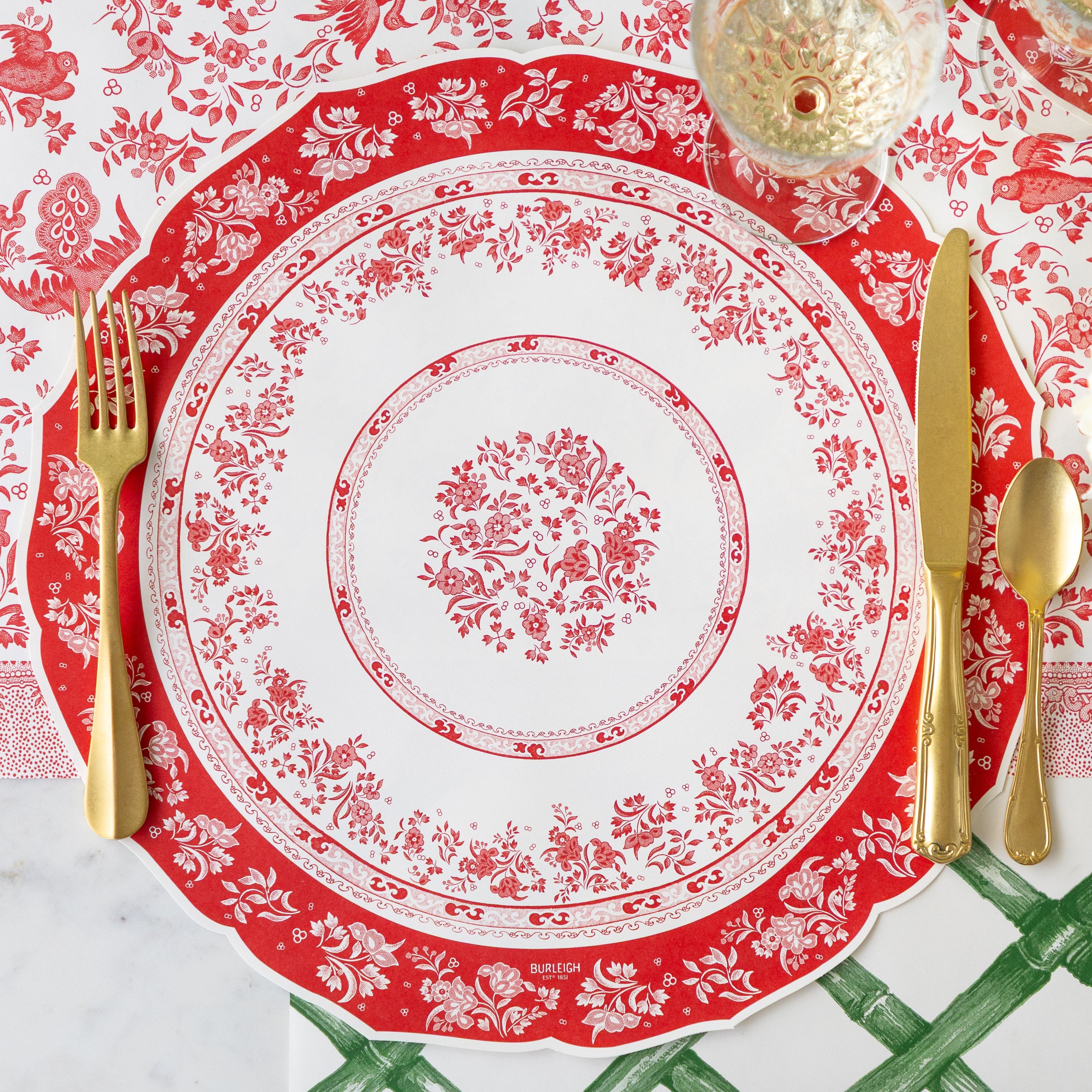 A place setting with no plates featuring the Die-cut Red Burleigh Placemat, gold flatware, a glass of wine, Red Regal Peacock Runner and Green Lattice Runner underneath.