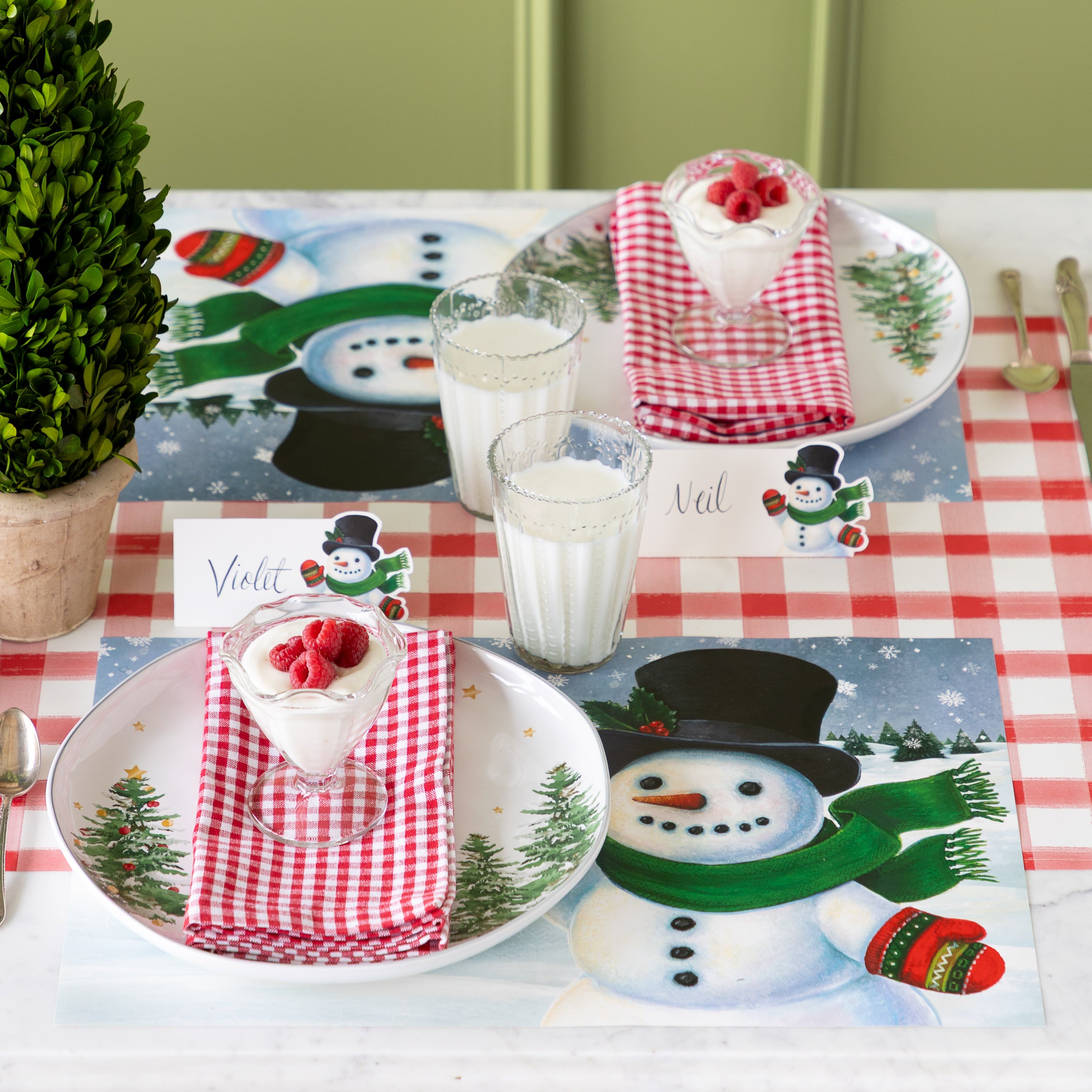 Two place settings with Jolly Snowman Placemats, red checkered napkins, parfait desserts topped with raspberries, and glasses of milk arranged on the Red Painted Check Runner.