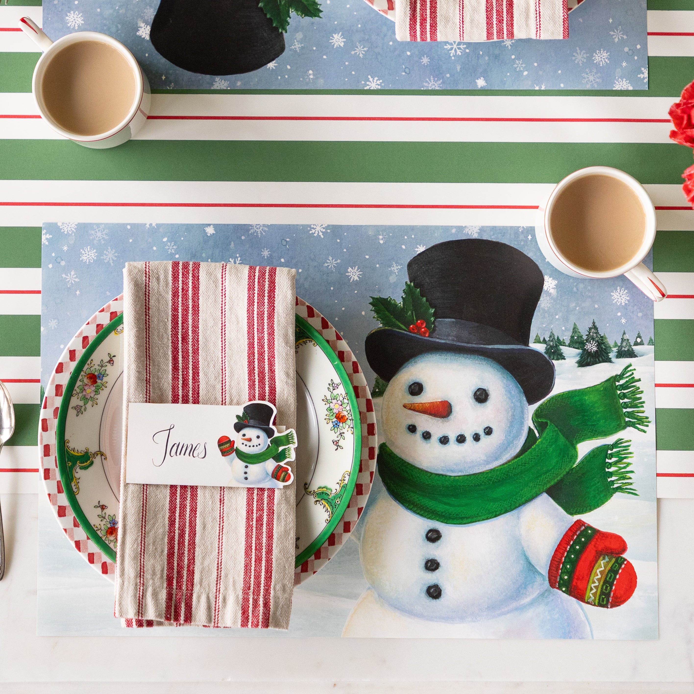 A festive place setting with the Jolly Snowman Placemat, Snowman Place Card reading "James" resting on a napkin on top of two dinner plates and Green & Red Awning Stripe runner underneath.
