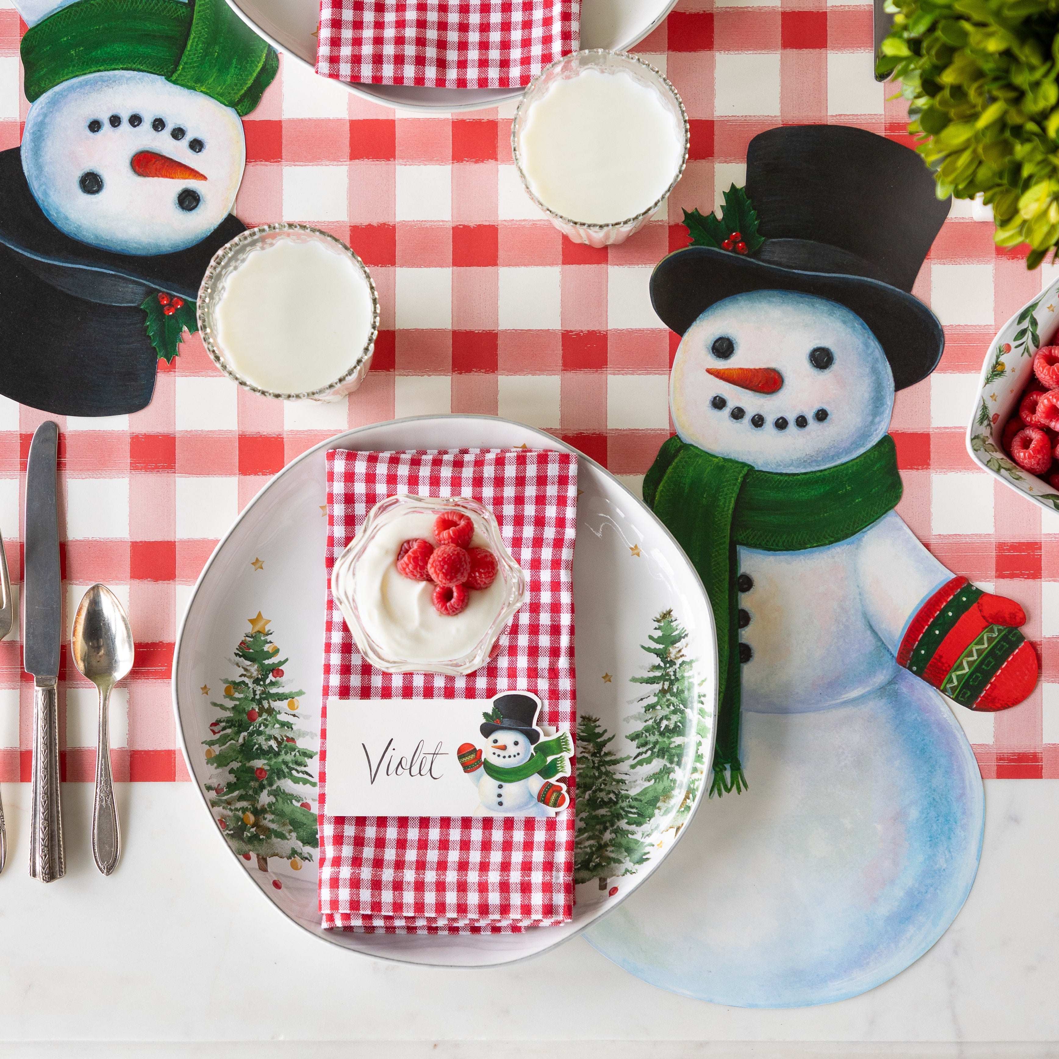 A festive table setting with the Die-cut Jolly Snowman Placemat, a festive tree plate, red checkered napkins with the Snowman place card on top and Red Painted Check Runner underneath.