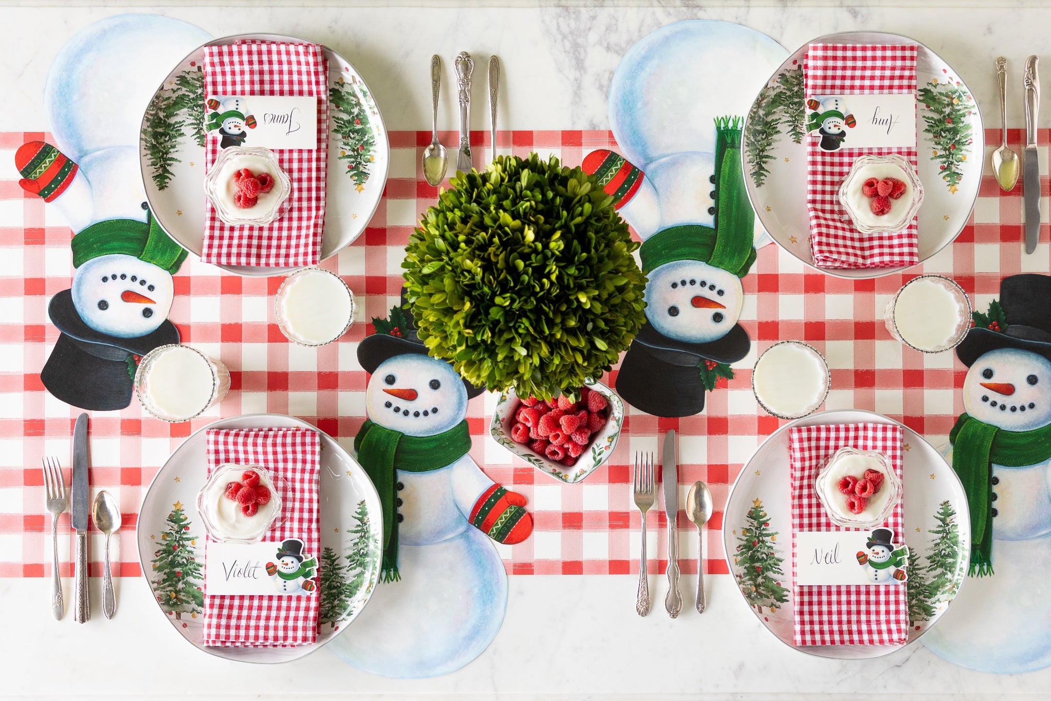A winter table setting featuring Die-cut Jolly Snowman Placemats, Snowman Place Cards, vintage dinnerware and topiary on the Red Painted Check Runner.
