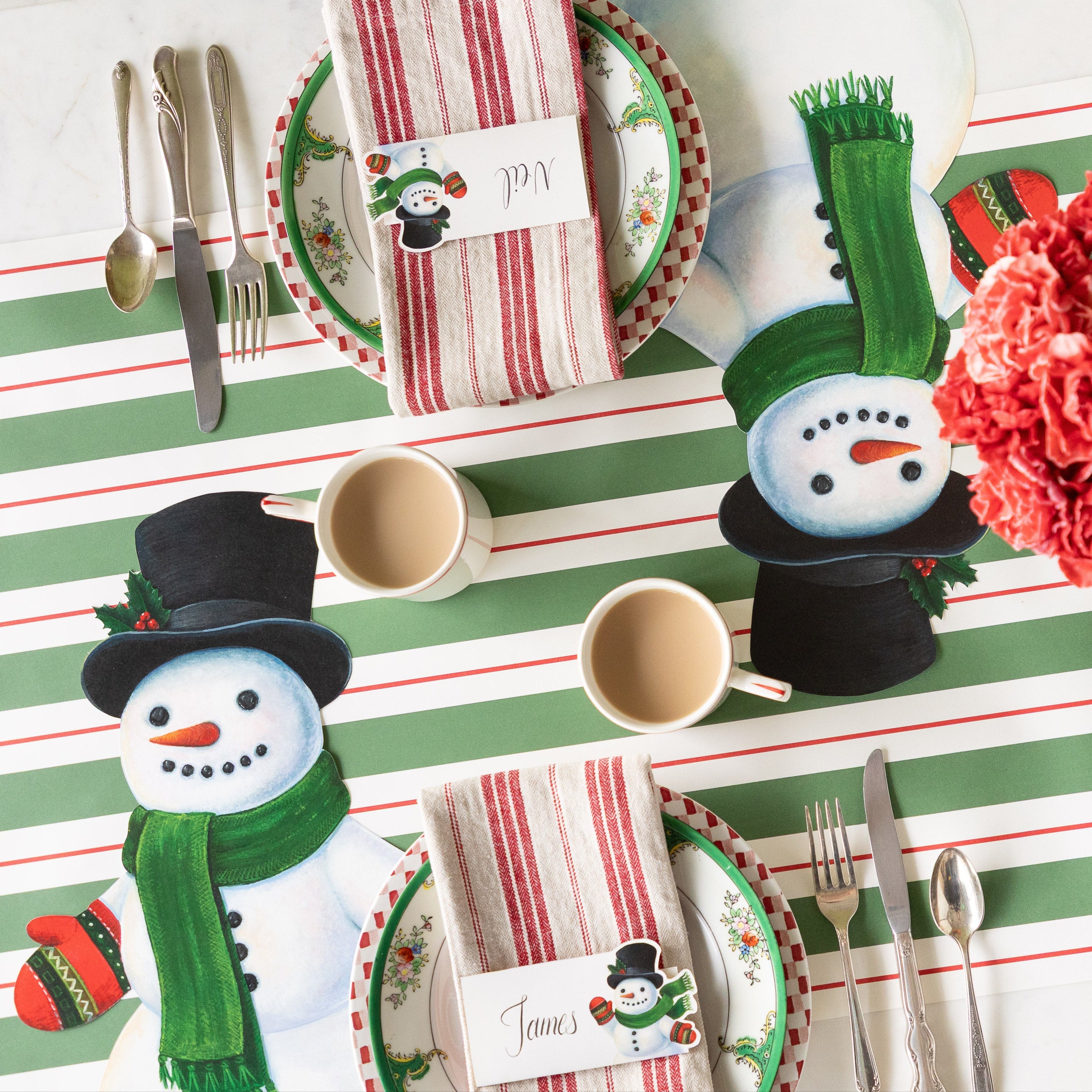 A festive table setting with the Die-cut Jolly Snowman Placemat, cups of coffee, red striped napkins with the Jolly Snowman Place Card on top and Green & Red Awning Stripe Runner underneath.