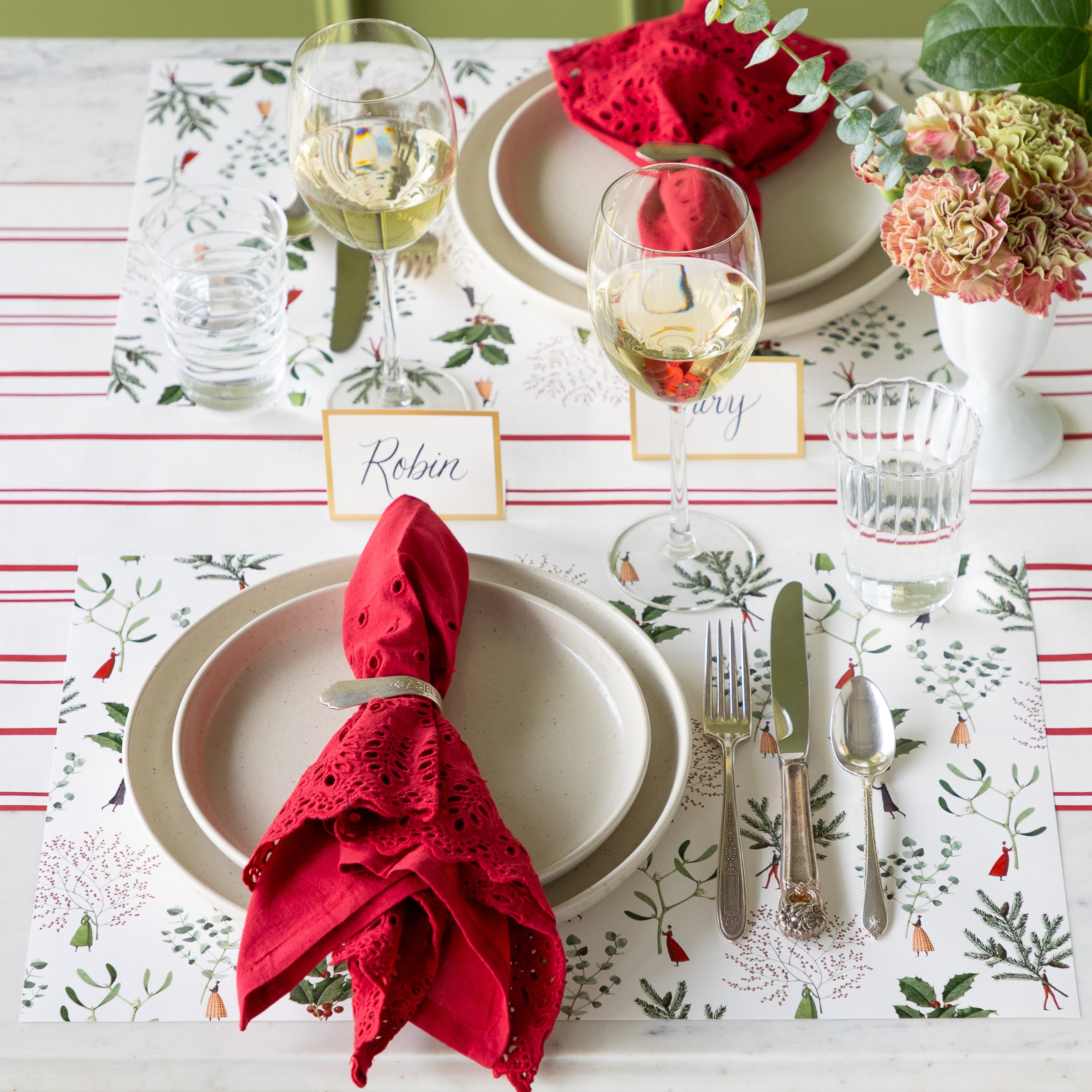 An elegant table setting featuring the Happy Christmas Placemats, dinner plates with a red lace napkin in a holder atop, vintage silverware, glasses of wine and water, Gold Frame Place Cards and Antique Red Stripe Runner underneath.