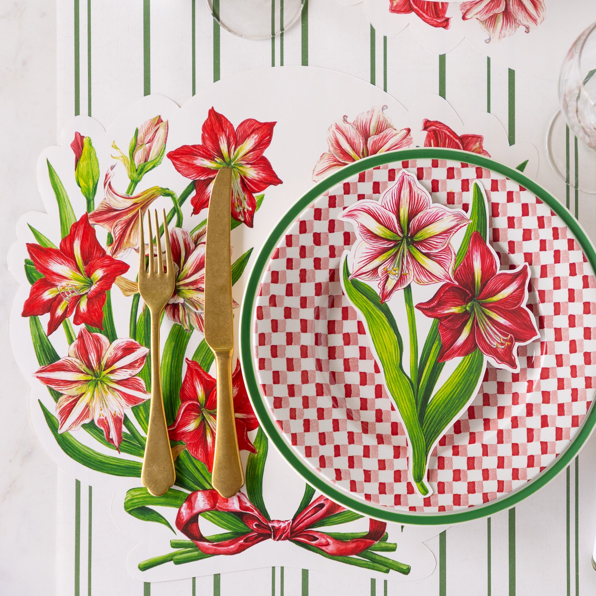 A place setting featuring the Die-cut Amaryllis Placemat with a green-rimmed and red-and white checkered plate and Amaryllis Table Accent atop, and gold flatware on the Antique Green Striped Runner.