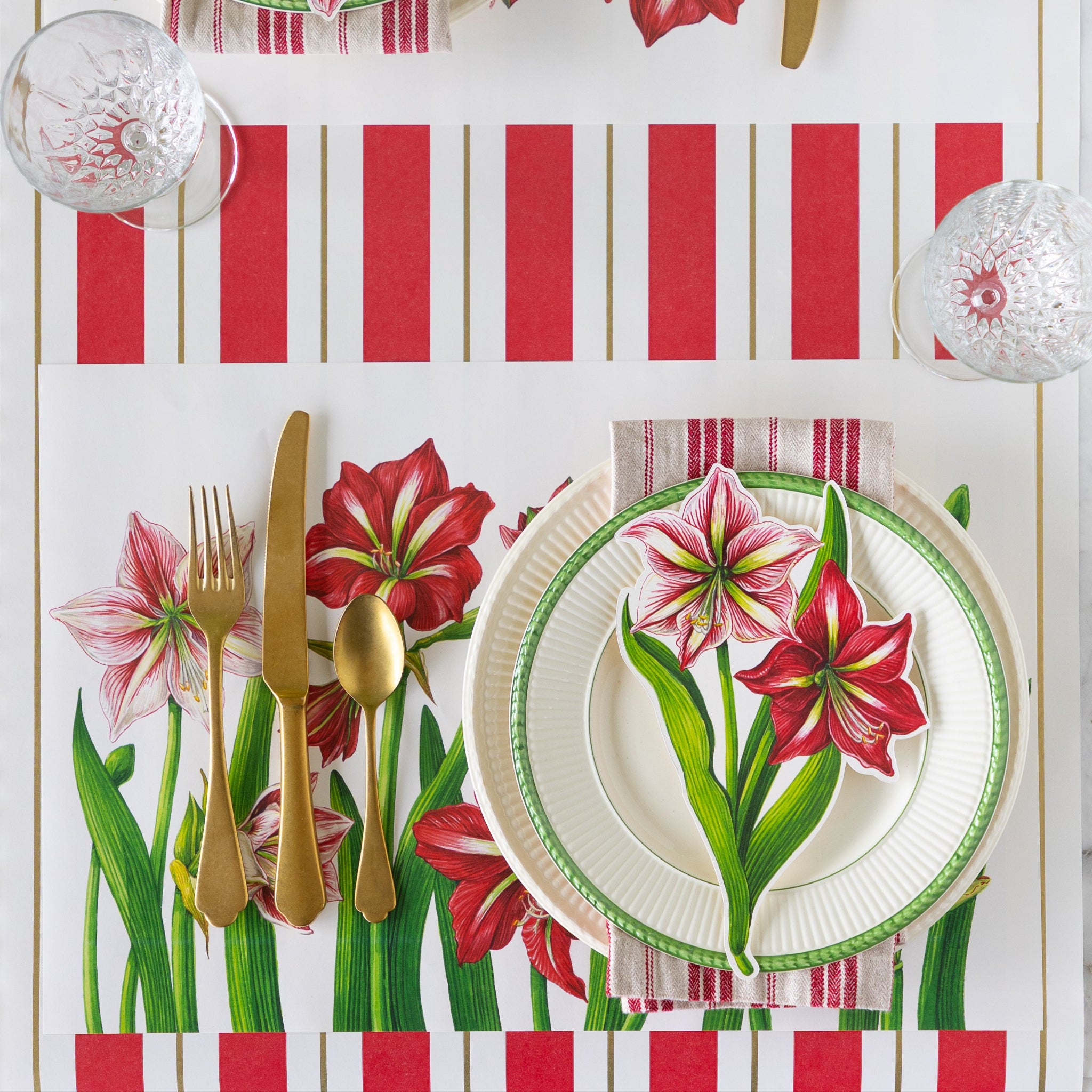 An elegant place setting featuring the Amaryllis Blooms Placemat, dinner plates, red-and-white striped napkin, Amaryllis Table Accent and gold flatware on the Red & Gold Awning Stripe Runner.