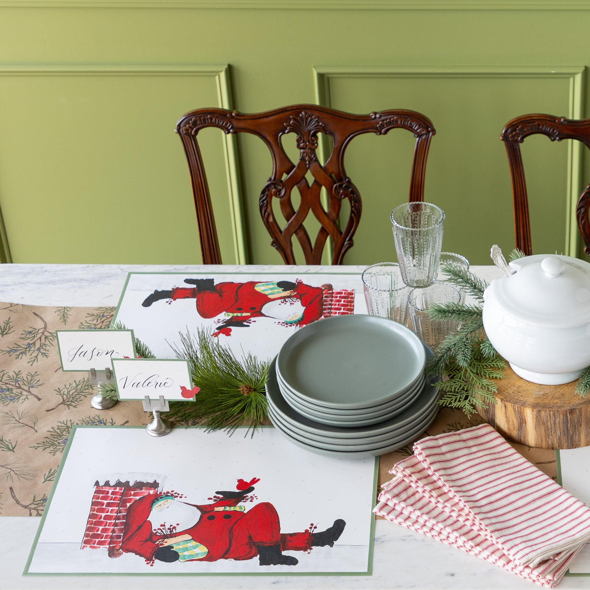 Set dining table Old St. Nick Chimney Placemats, red-and-white striped napkins, green plates, Red Bird Place Cards and woodland decor on the Juniper Sprigs on Kraft Runner.