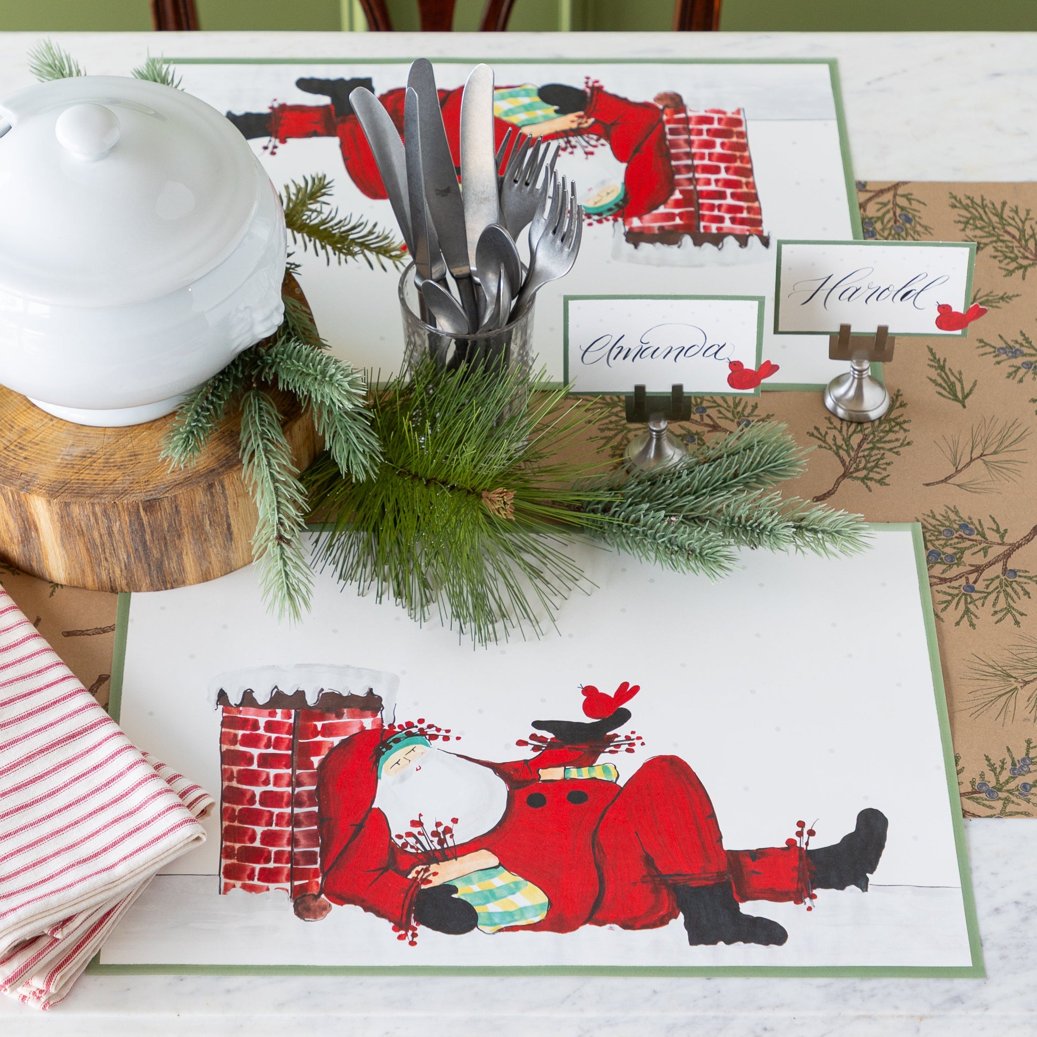 Christmas themed table setting featuring the Old St. Nick Chimney Placemats, red-and-white striped napkins, vintage silverware, Red Cardinal Place Cards and woodland decor on the Juniper Sprigs on Kraft Runner.