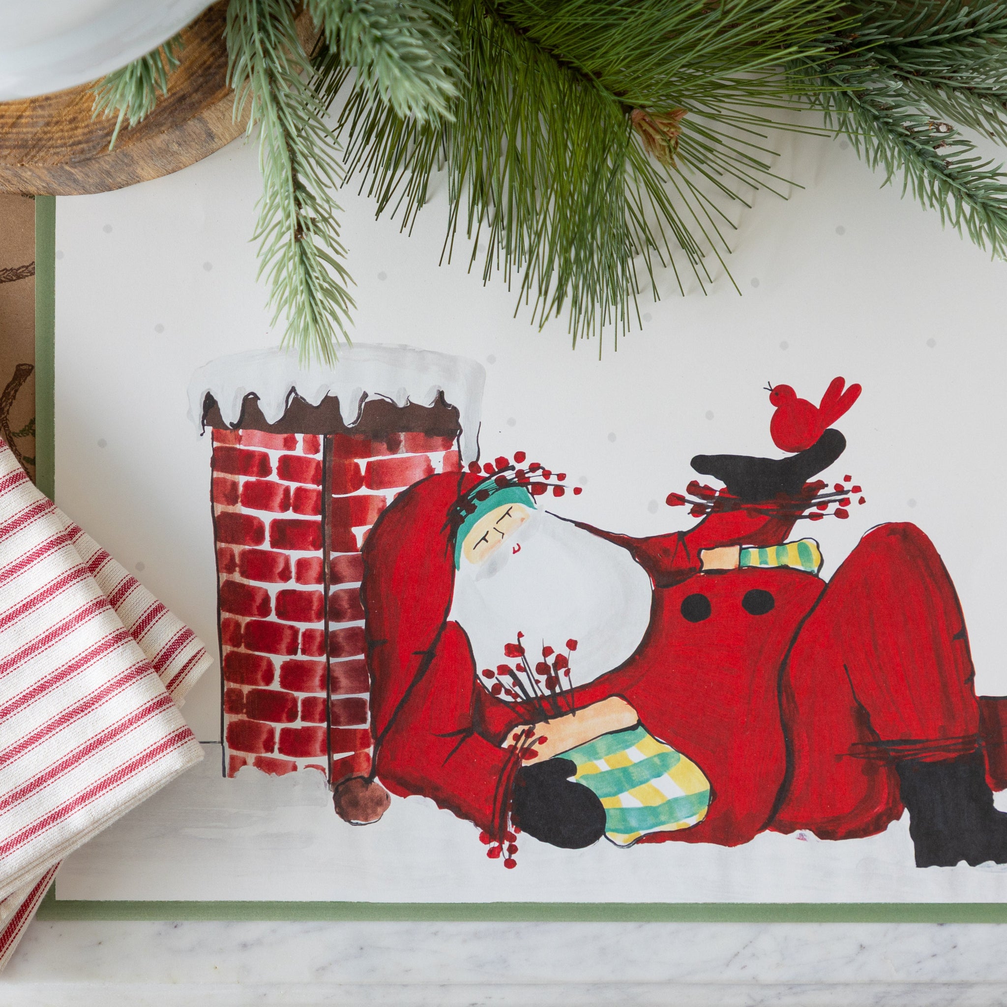 Christmas themed table setting featuring the Old St. Nick Chimney Placemats and red-and-white striped napkins on the Juniper Sprigs on Kraft Runner.