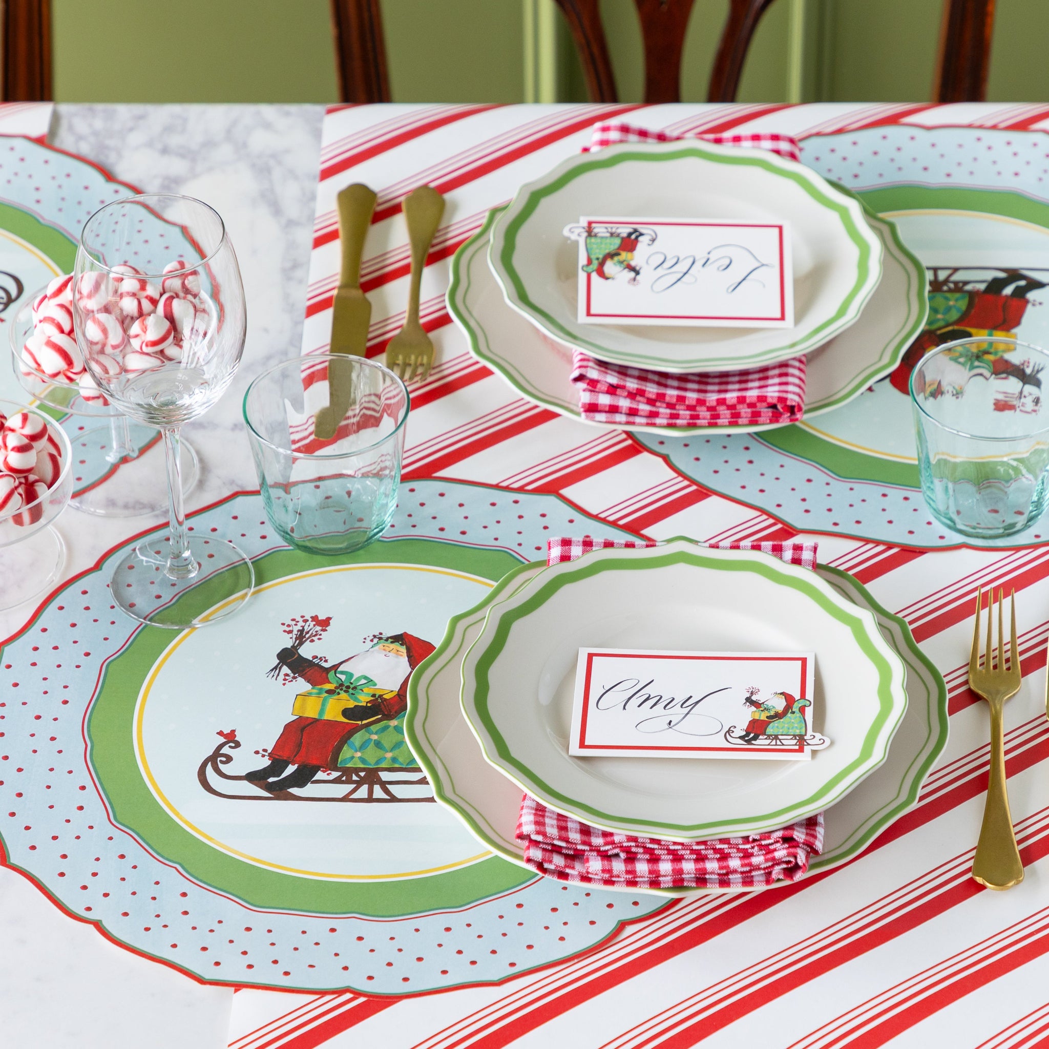 Christmas-themed table setting with Green Icing on the Cake plates, red-and-white checkered napkins, gold cutlery, and Die-cut Old St. Nick Sleigh Placemat on the Peppermint Stripe Runner.