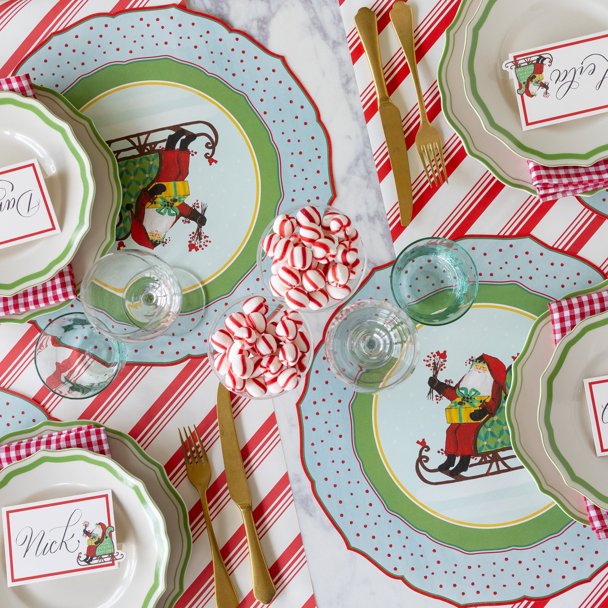 Christmas-themed table setting with Green Icing on the Cake plates, red-and-white checkered napkins, gold cutlery, cups of peppermints and Die-cut Old St. Nick Sleigh Placemat on the Peppermint Stripe Runner.