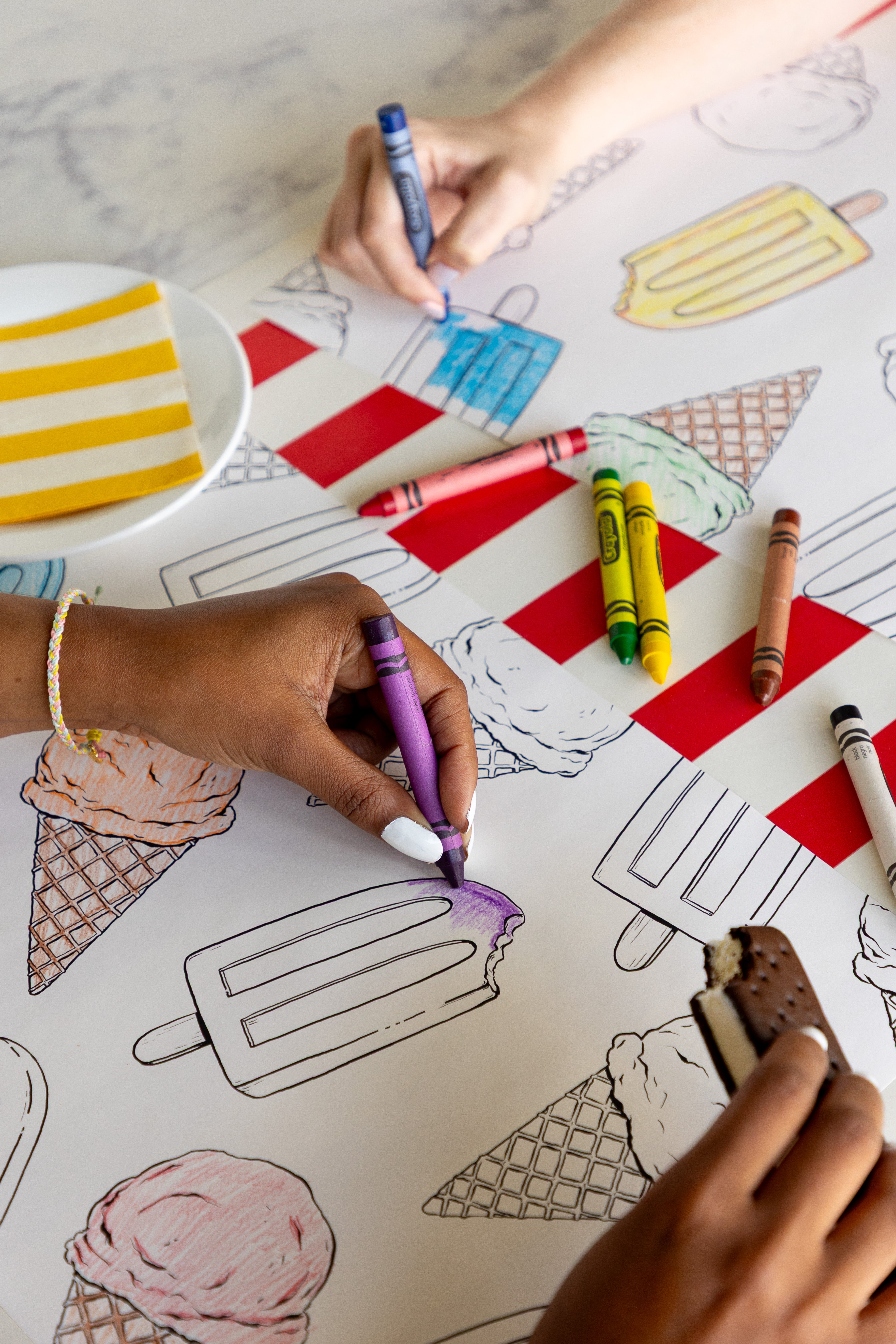Two people coloring the Coloring Summer Treats Placemats with crayons on a table with the Red Classic Stripe Runner.