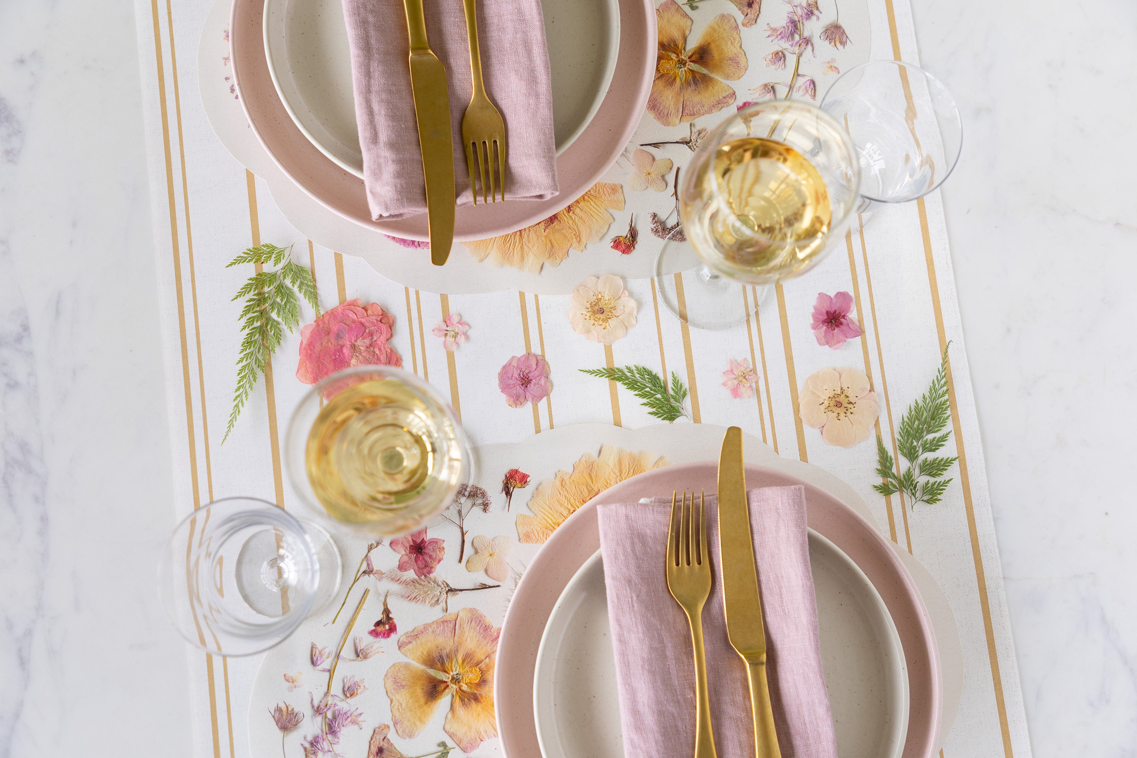 Table setting with floral placemats, pink plates, gold cutlery, and white wine glasses.