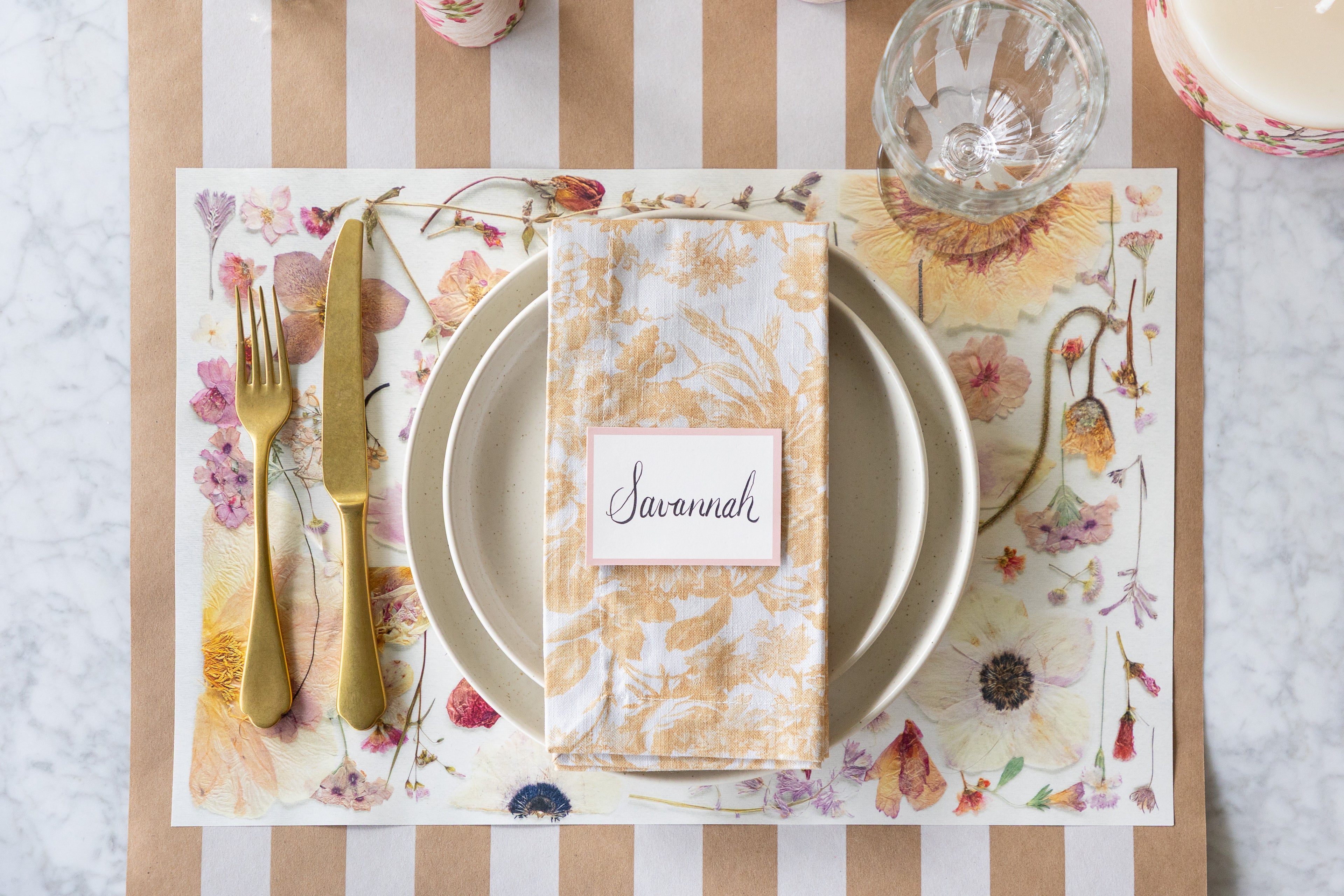 Table setting with floral placemat, plates, cutlery, and name card on a striped tablecloth.