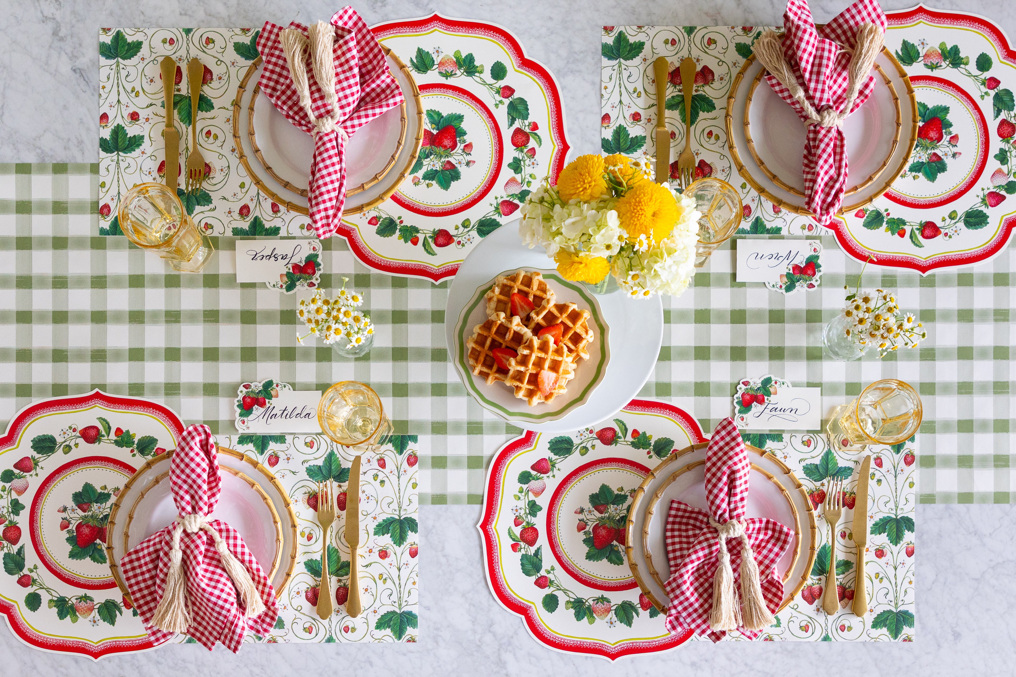 Decorative table setting with waffles, flowers, Die-cut Strawberry China and Strawberry Fields Placemats, Strawberries Place Cards, red checkered napkins and gold cutlery on the Moss Painted Check Runner.