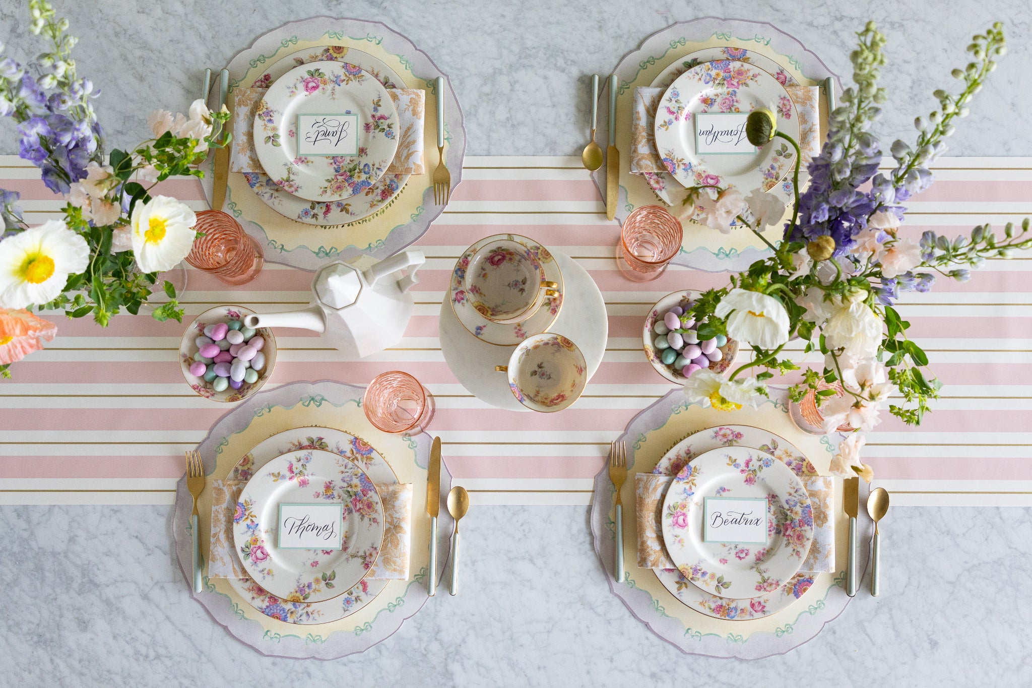 Easter table setting with flowers, floral plates, pastel glasses, Easter eggs and Die-cut Spring Meadow Placemats on the Pink & Gold Awning Stripe Runner.