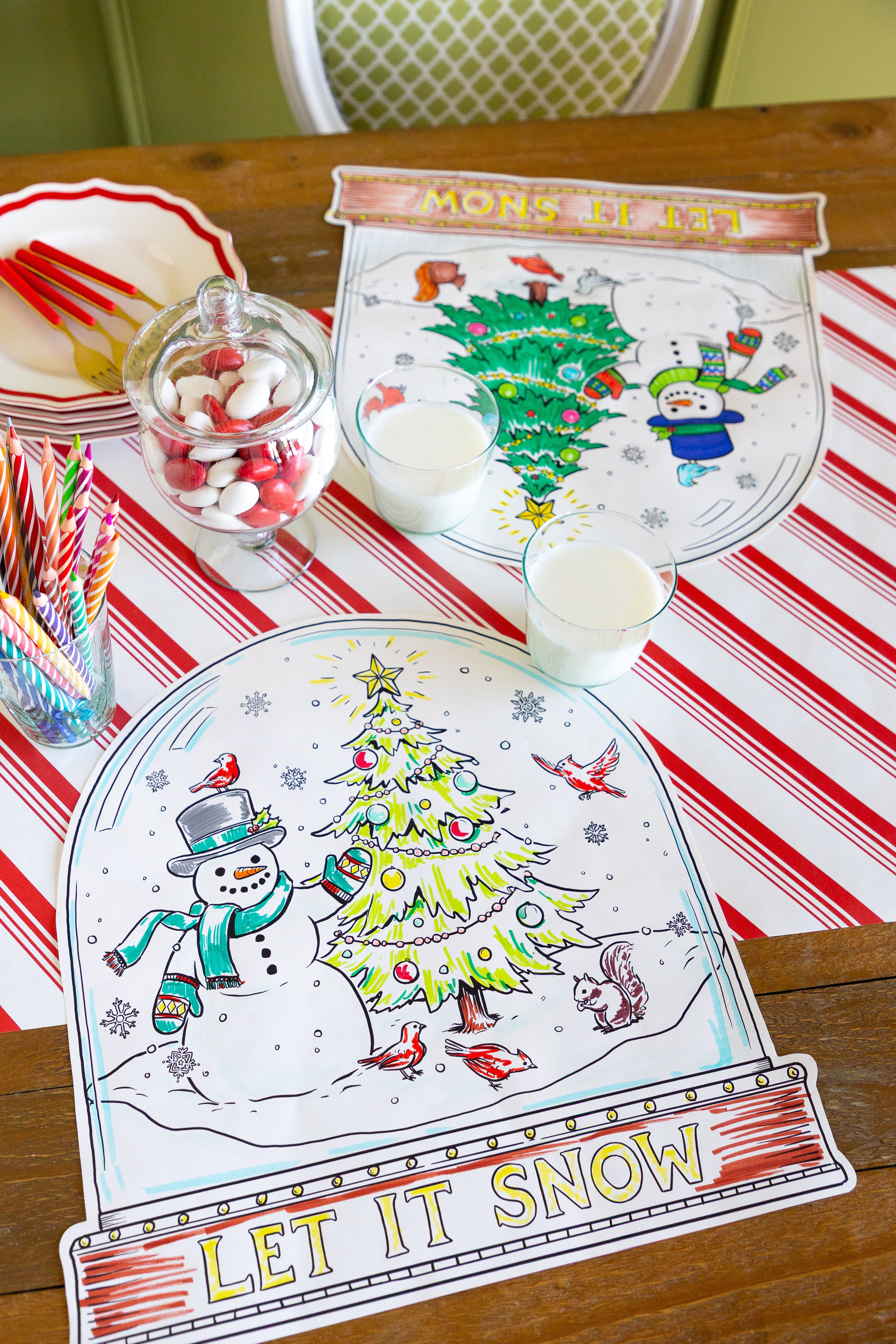 Table with two partially colored in Die-cut Snow Globe Coloring Placemats on a Candy Stripe Runner with glasses of milk and candy on the table.