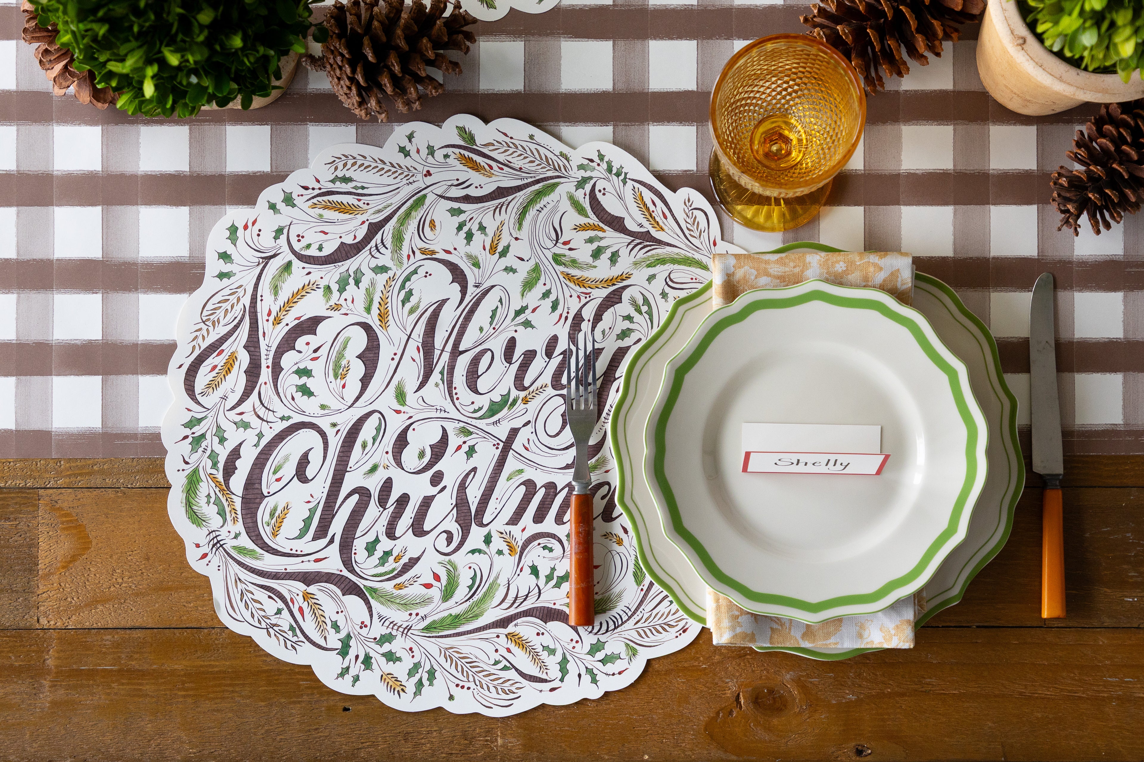 Christmas table setting with a decorative placemat, plates, and cutlery on a brown painted check runner.