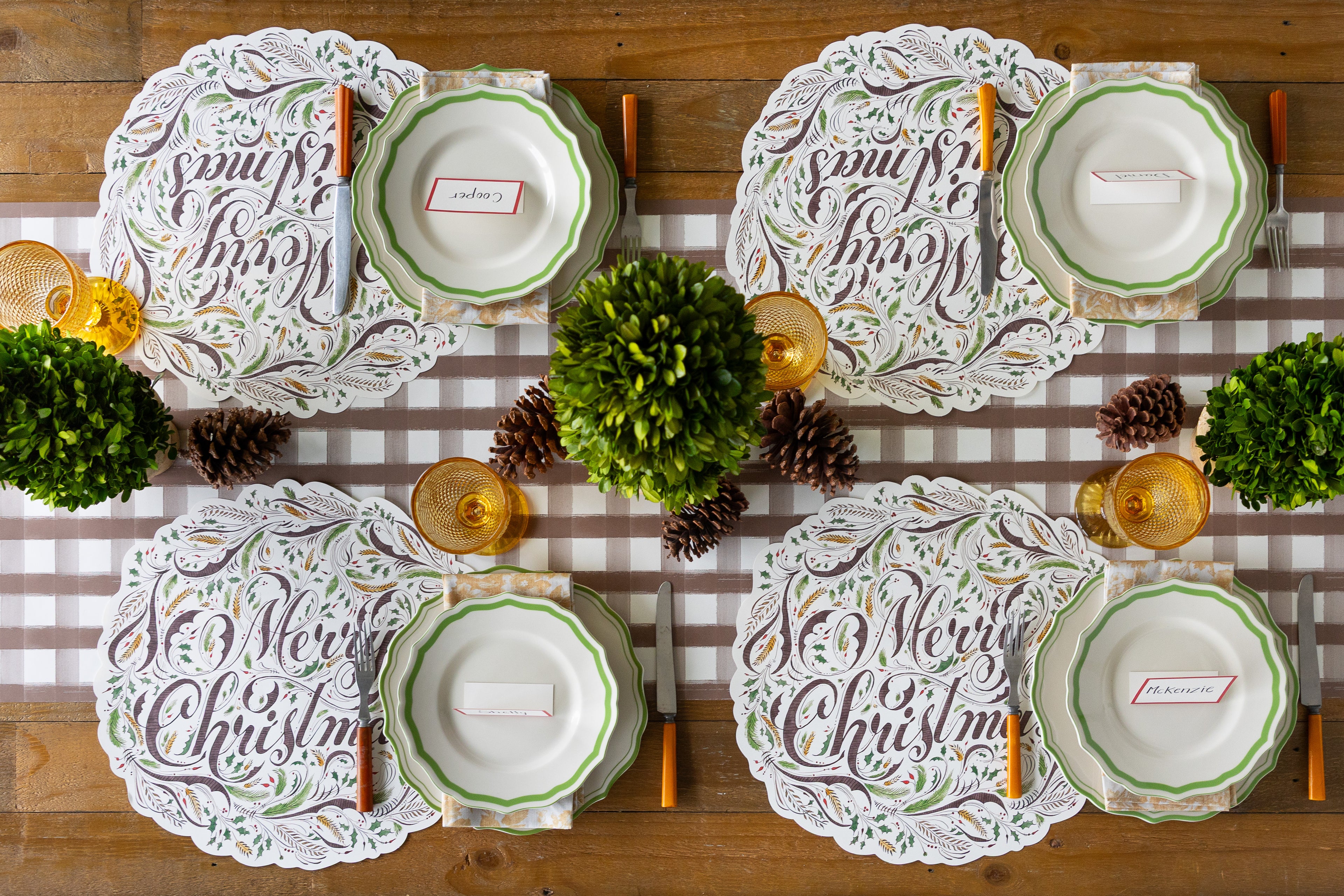 A Christmas table setting with the brown painted check runner down a wood table with four Die-cut Merry Christmas Placemats on it. There are plates, cutlery and glassware to match the theme.