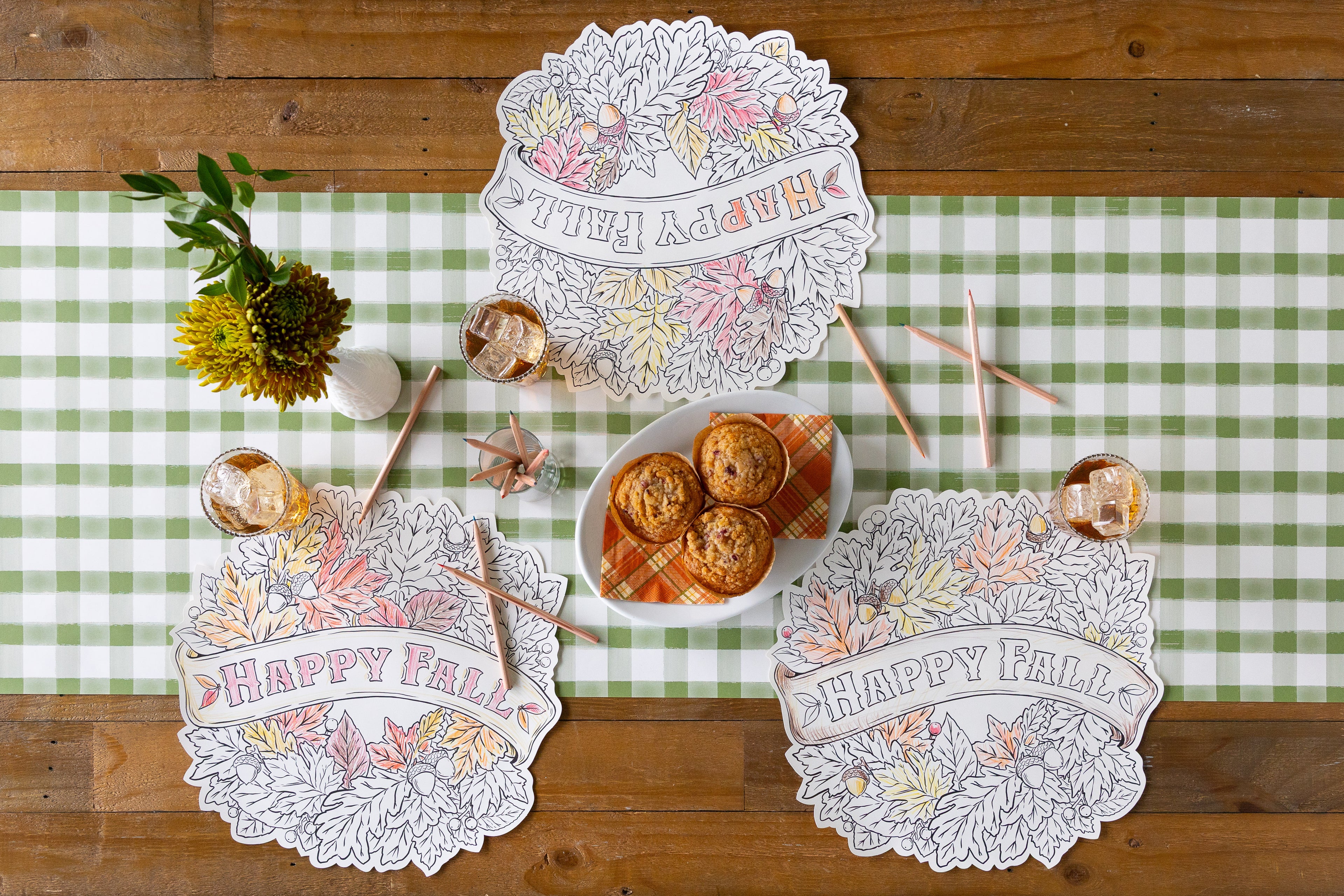 Tablescape with three Die-cut Happy Fall Coloring Placemats on top of a green check table runner with color pencils laid out with flowers and snacks.