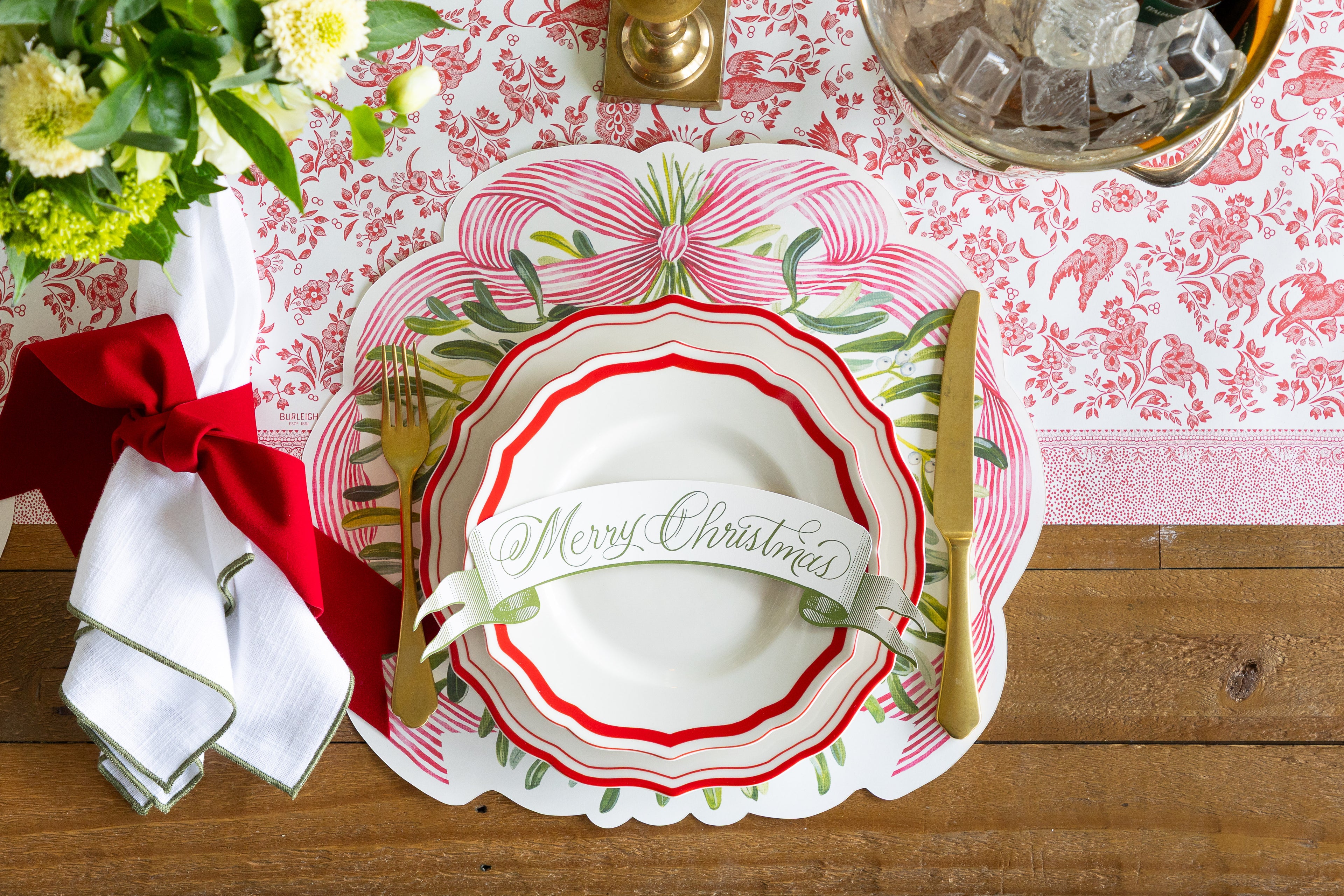 Christmas-themed table setting with decorative plates, cutlery, and a red ribbon on a wooden table.