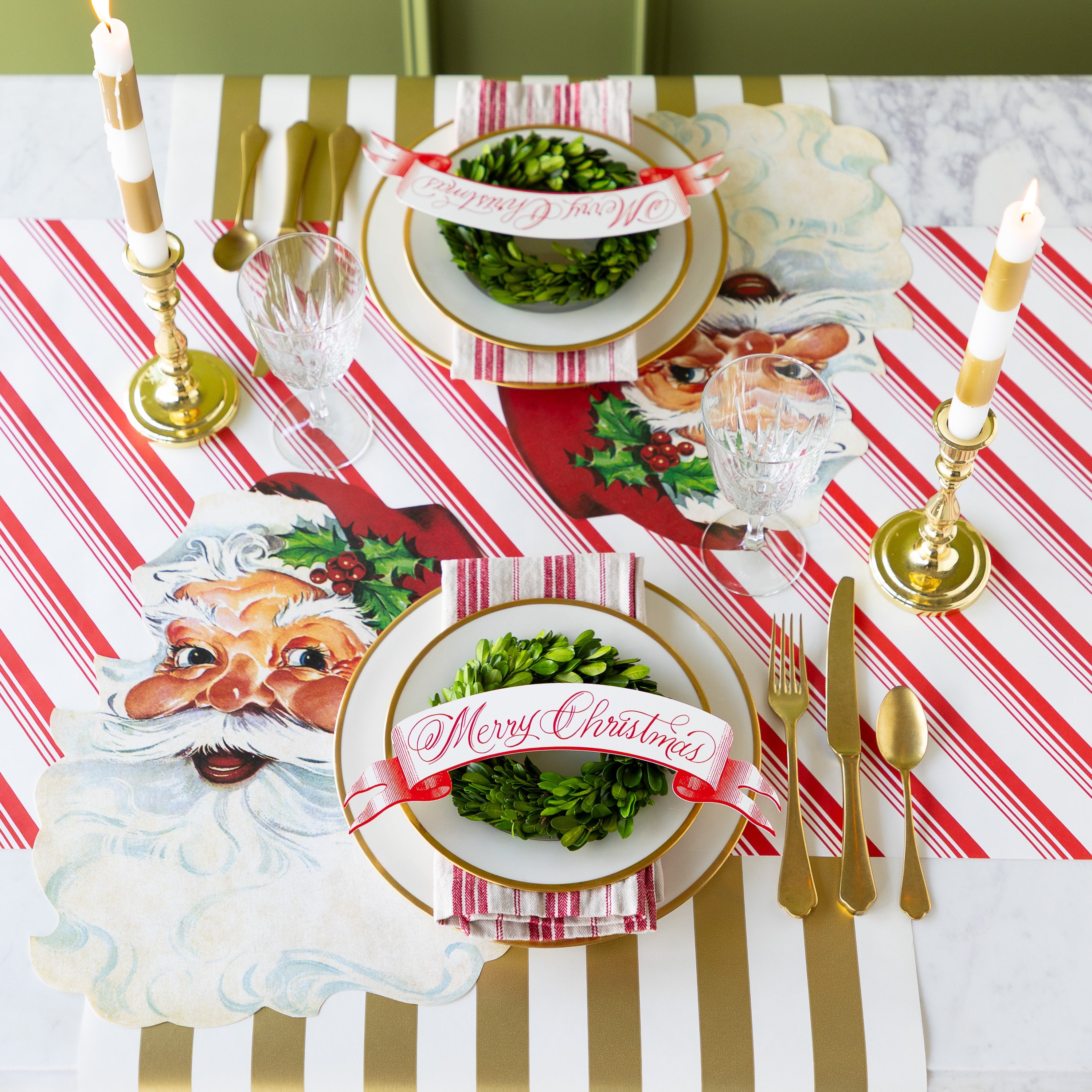 Christmas-themed table setting with Die-cut Santa Claus Placemats, Merry Christmas Red Banner Table Accents, candles, topiary wreaths and gold flatware on the Peppermint Stripe and Gold Classic Stripe Runners.