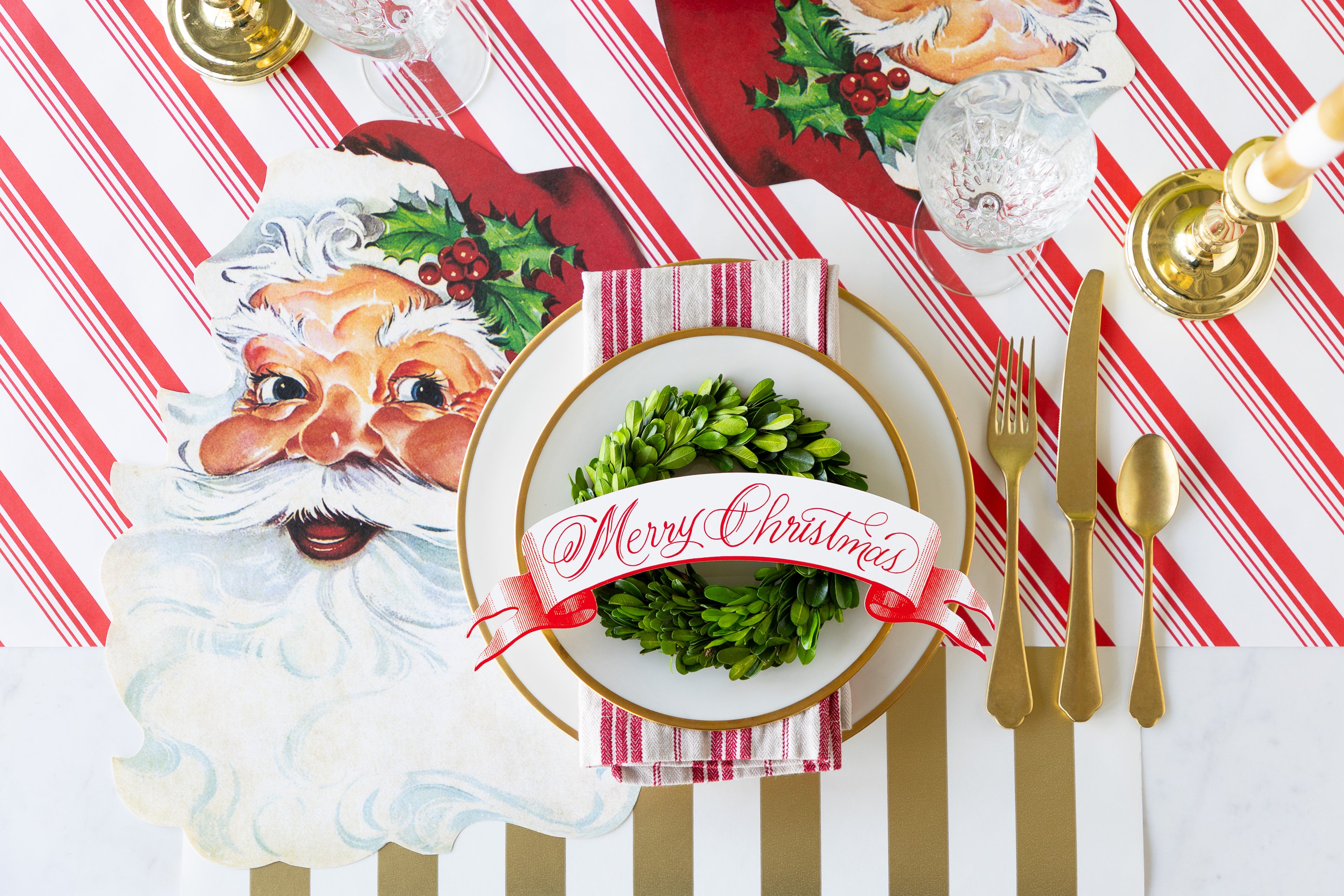 Christmas-themed table setting with Santa Claus placemat, gold cutlery, and red and white striped tablecloth.