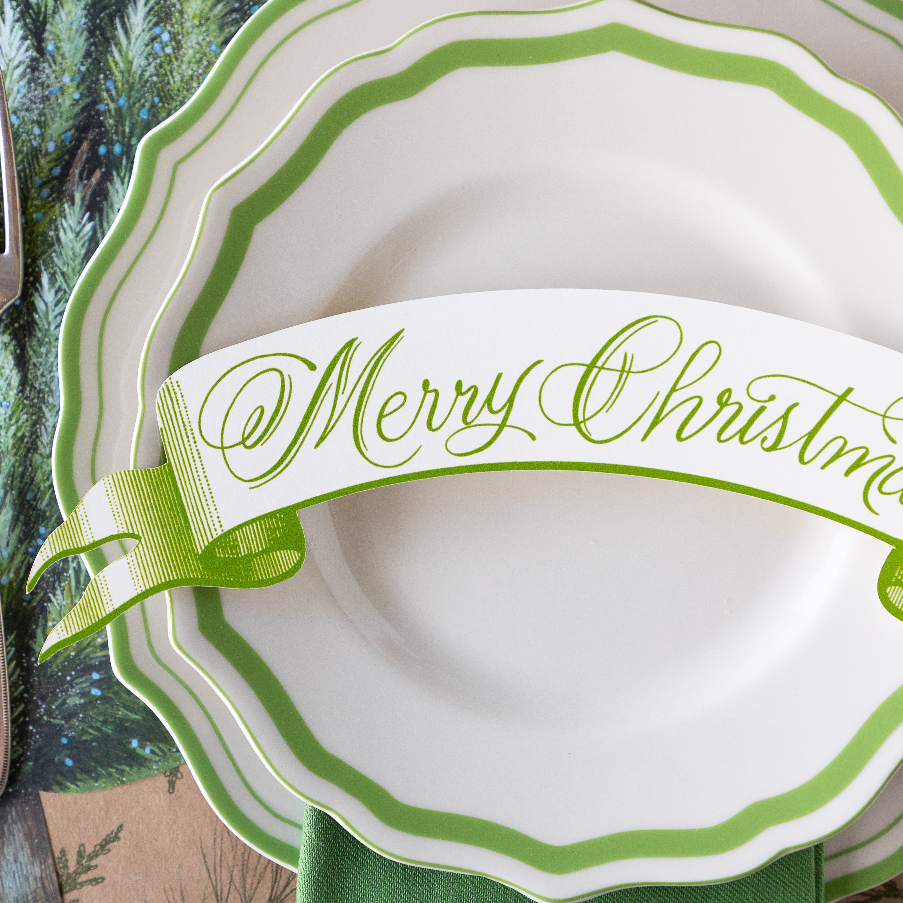 A close up of the Moss Merry Christmas Banner Table Accent atop green-and-white rimmed plates.