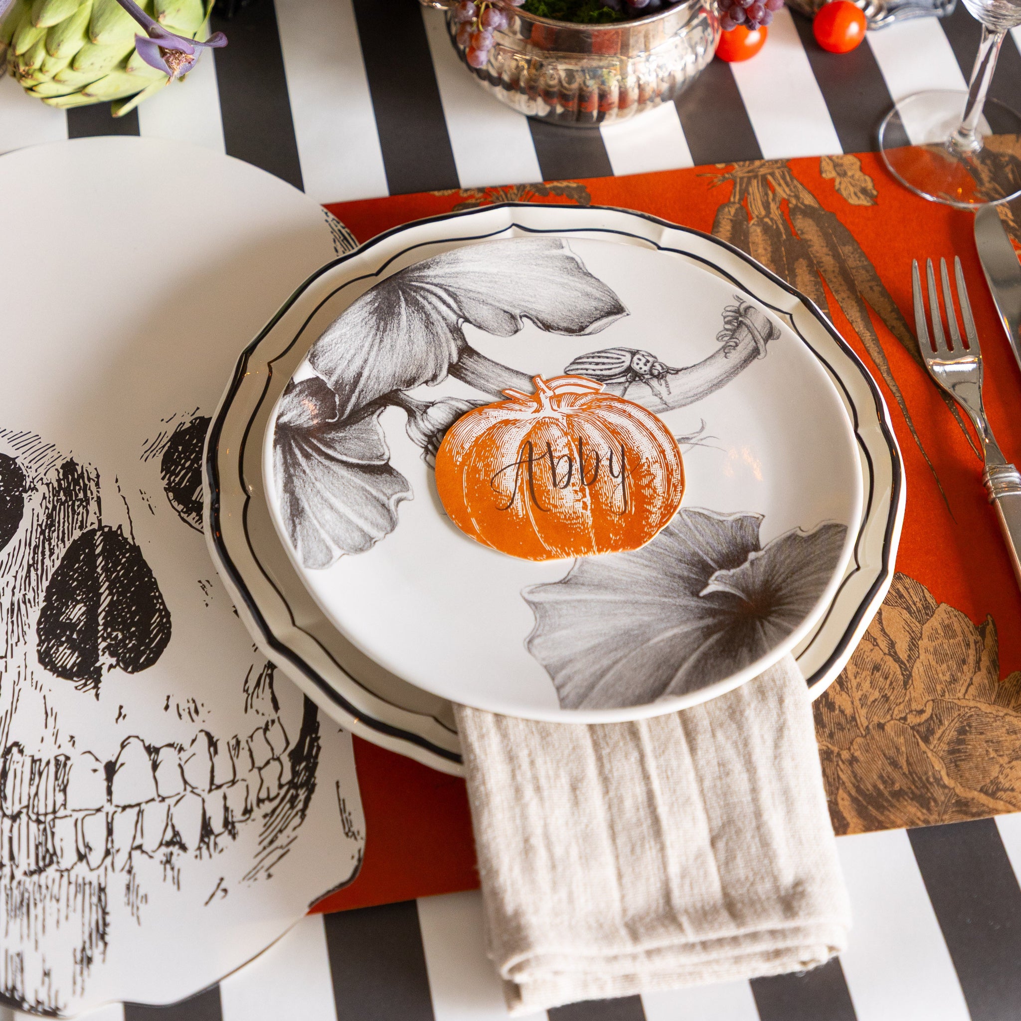 A Halloween themed place setting with the Die-cut Skull Placemat, decorative plates, Pumpkin Place Card and Harvest Vegetable Persimmon Placemat on the Black Classic Stripe Runner.