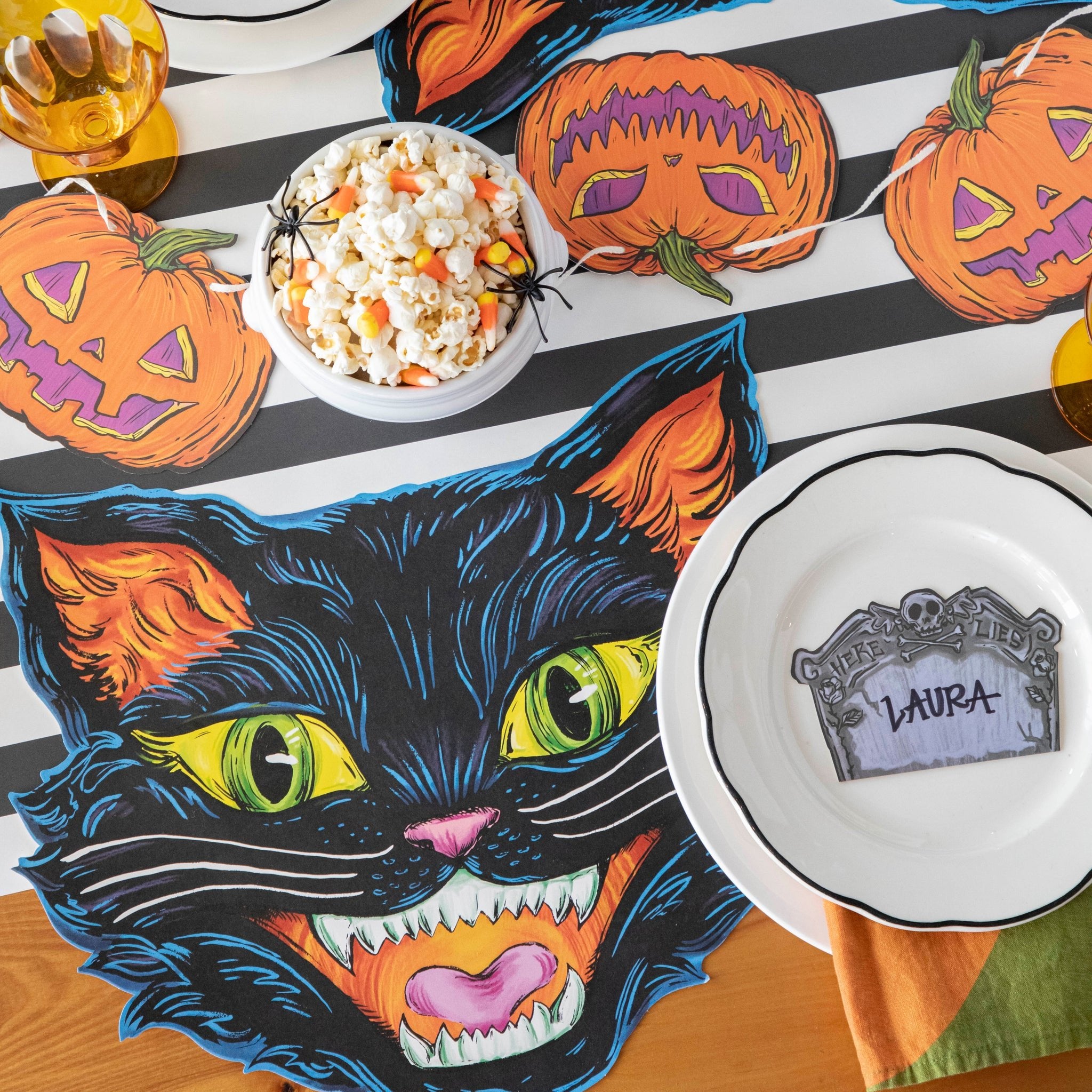 A spooky soiree table setting featuring the Black Classic Stripe Runner, adorned with pumpkin bunting, a bowl of popcorn and candy corn, and a white plate with the Tombstone Place Card labeled "Laura".
