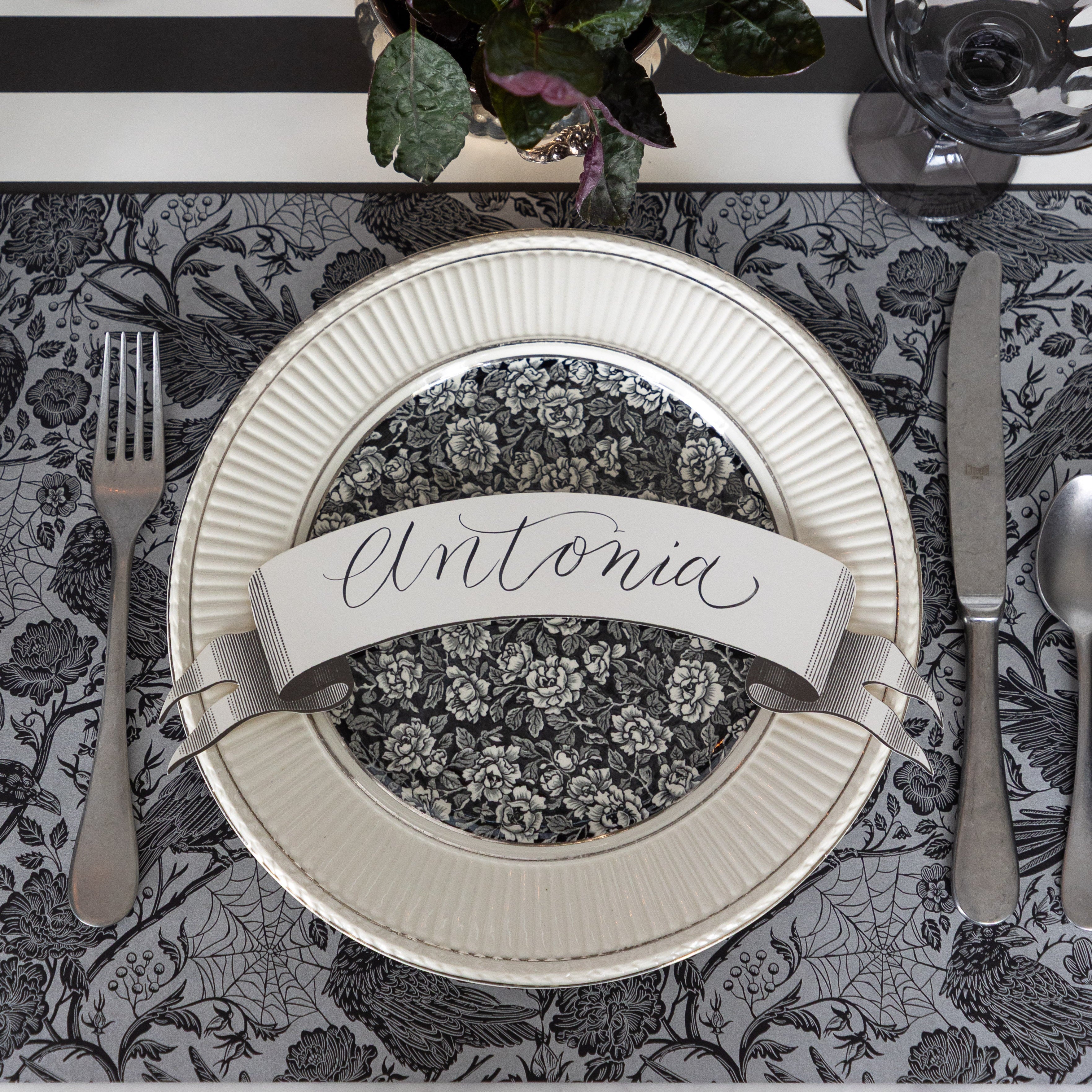 A spooky place setting featuring the Raven Toile Placemat with silver flatware, a dinner and salad plate with the Classic Black Banner Table Accent with "Antonia" written on it on top and Black Classic Stripe Runner underneath.