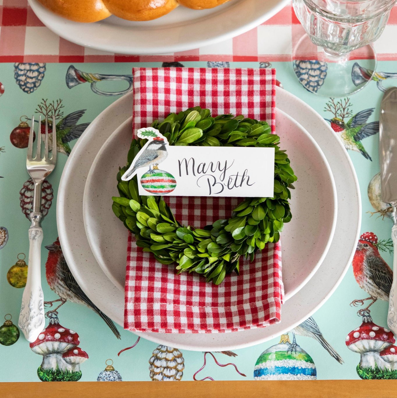 The Christmas Chickadee Place Card with "Richard" written on it, on top of a boxwood wreath and red painted check napkin, resting on dinner plates.