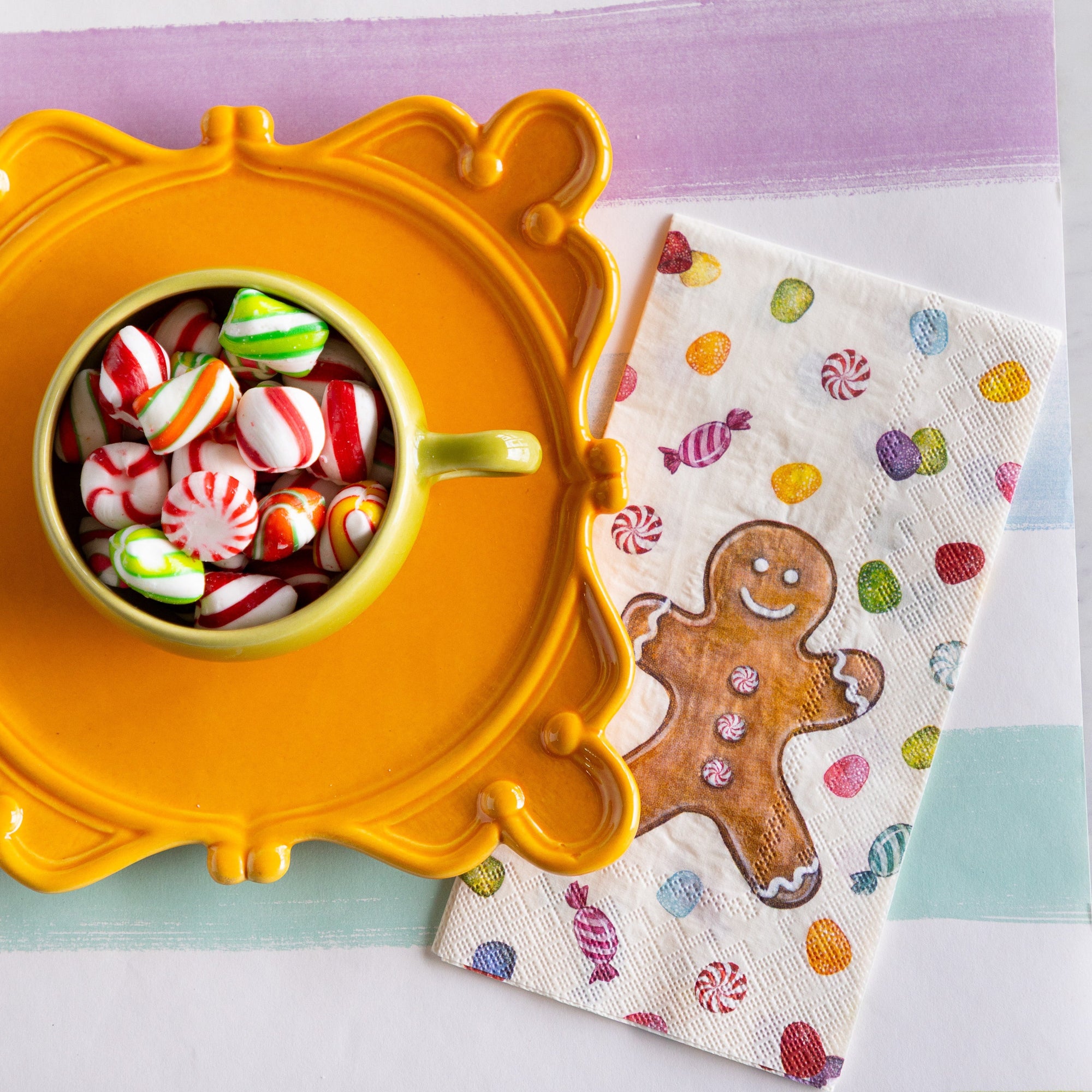 The Gingerbread Guest Napkin next to a decorative pedestal with a mug of candy atop, on the Sorbet Painted Stripe Runner.
