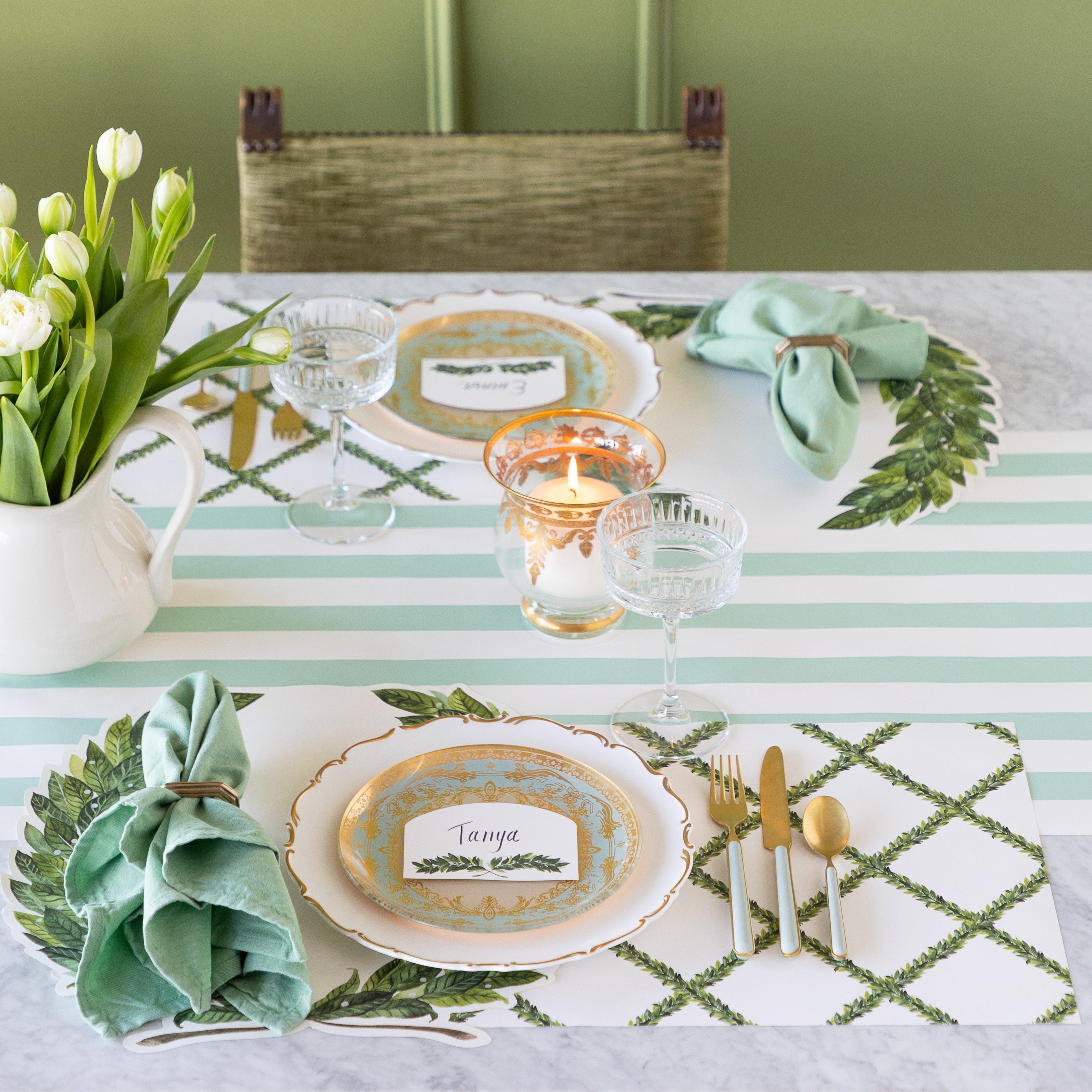 A candle lit table set for two with green and white decor, a vase of white tulips, Die-cut Laurel Wreath Placemats and Laurel Lattice Placemats with vintage dinnerware in between and the Laurel Place card atop, and Seafoam Classic Stripe Runner underneath.