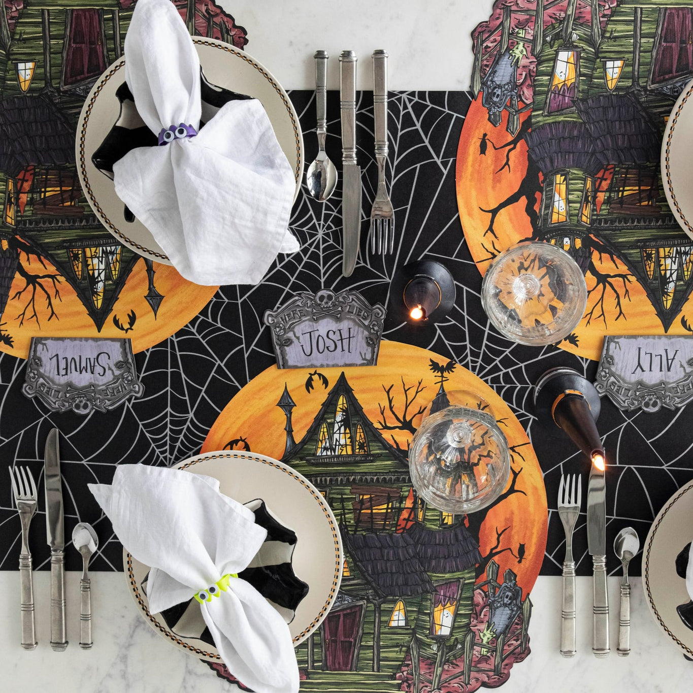 A spooky table setting featuring the Die-cut Haunted House Placemat, Tombstone Place Cards, lit witch hat candles, fake monster teeth around napkins, and Spiderweb Runner underneath.